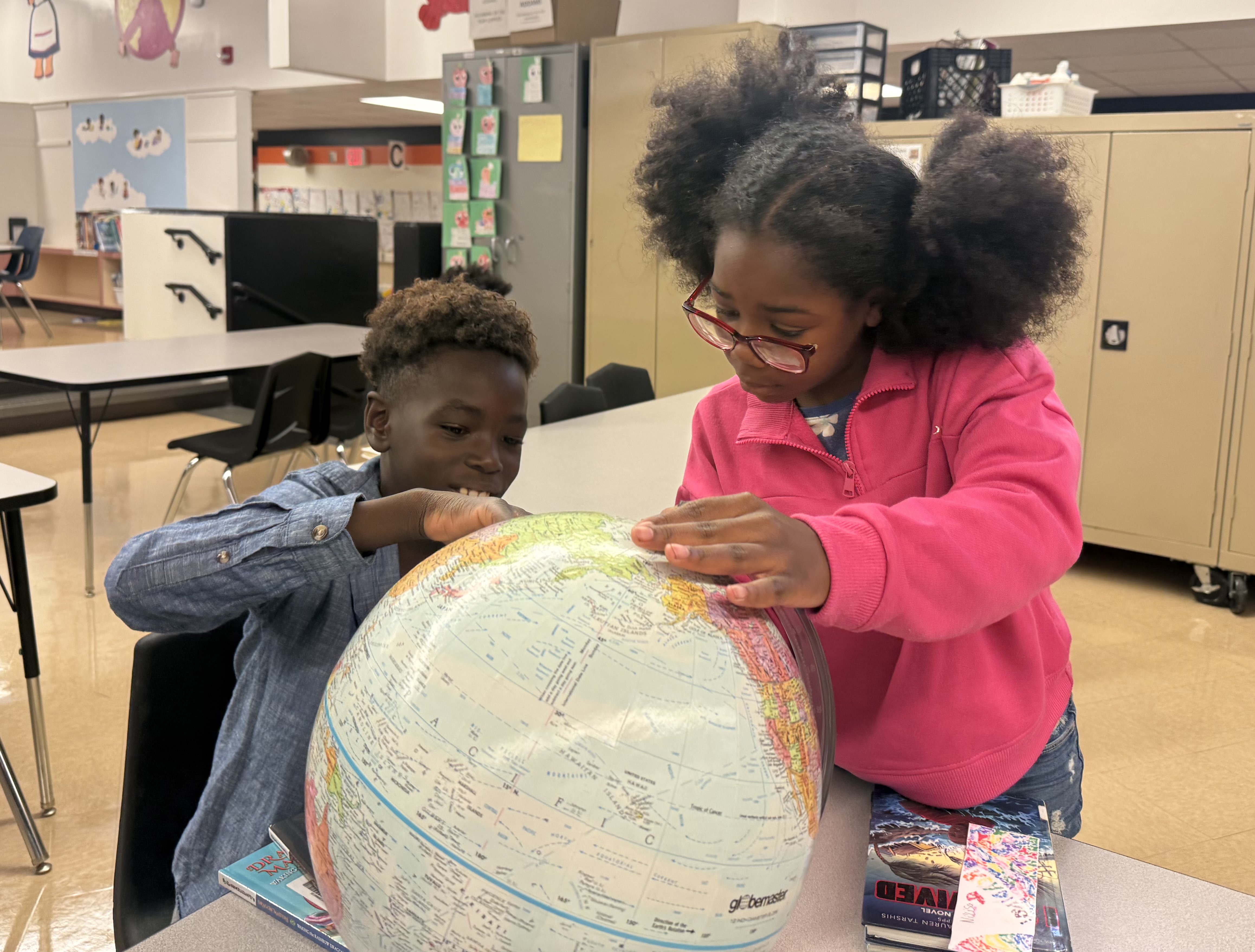 Two children, one with glasses, examining a globe on a table in a classroom with chairs.