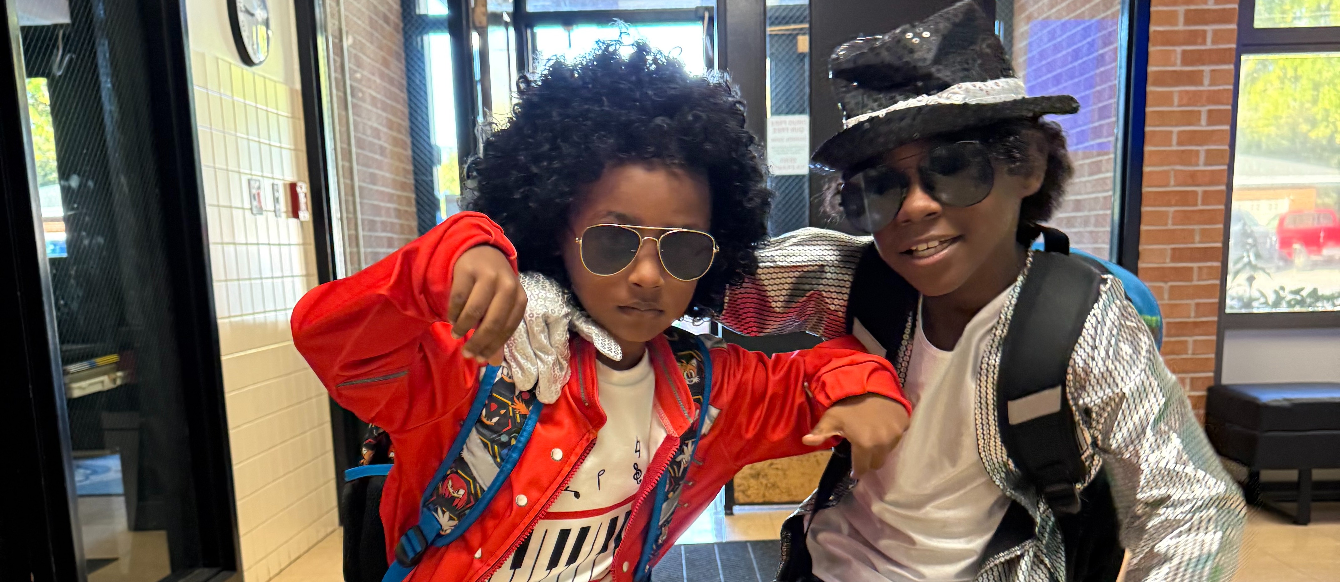 Two children in a hallway, one wearing a red jacket, the other in silver, both dressed in colorful costumes.