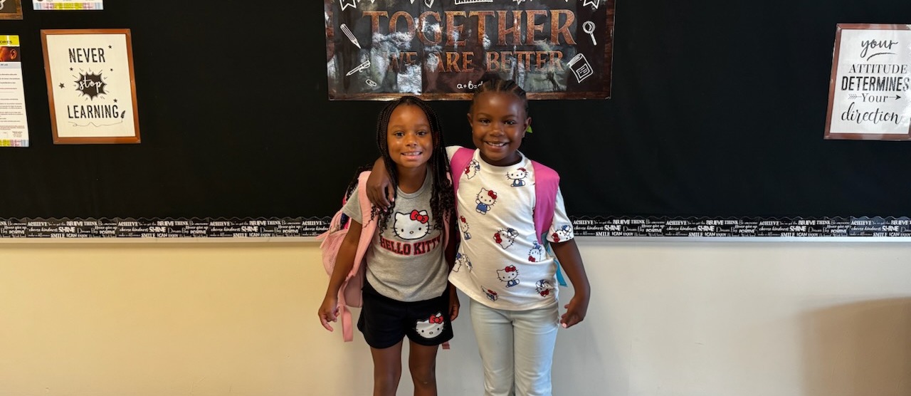 Two children standing before a classroom wall with a sign that reads "Welcome Together We Are Better."