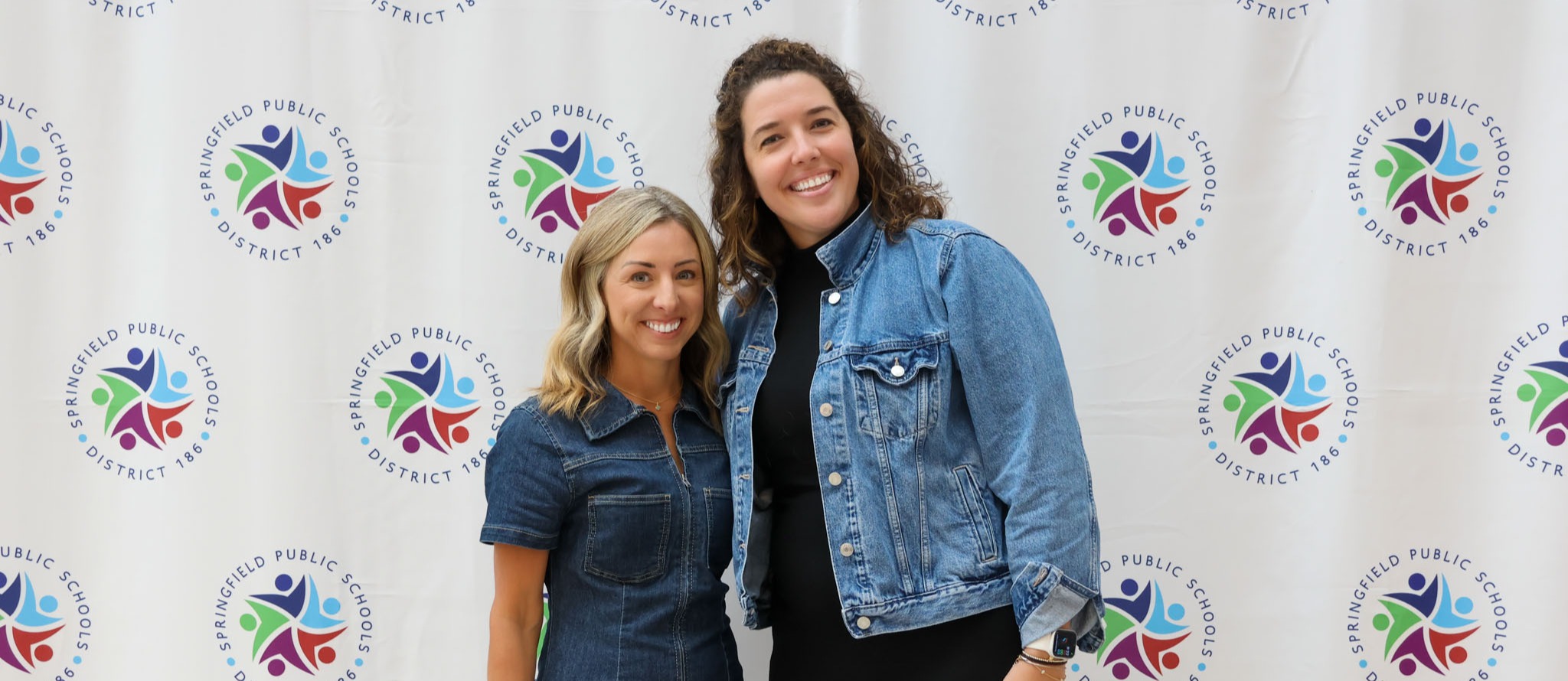 Two women, one in a denim dress and the other in a denim jacket, stand before a colorful backdrop.