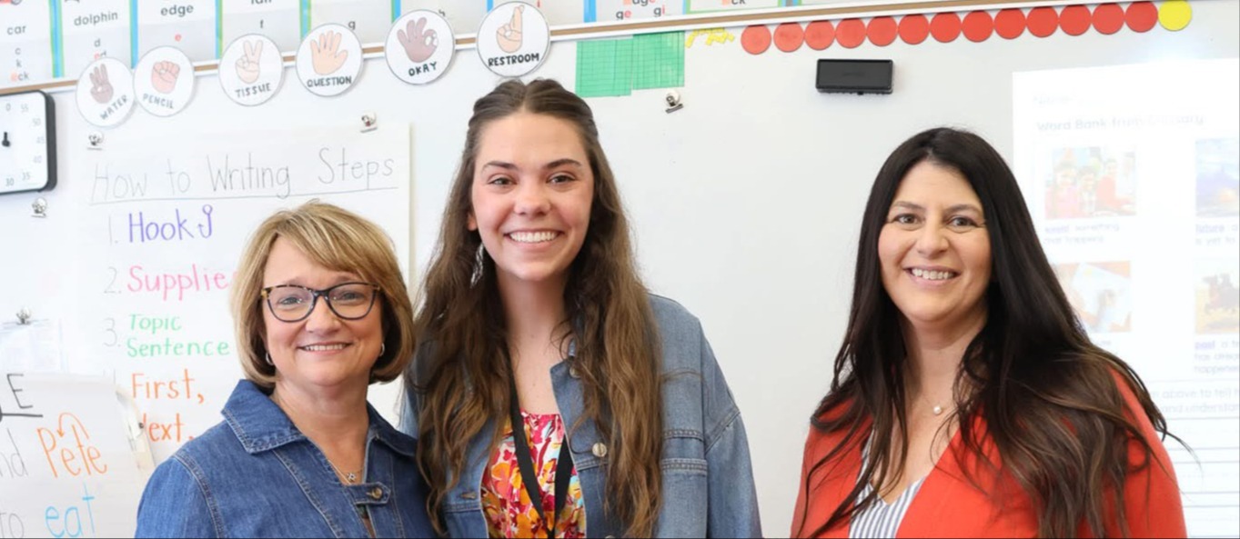 Three women stand in a classroom. One holds a certificate, another wears glasses, and the third has long hair. A colorful carpet is on the floor.