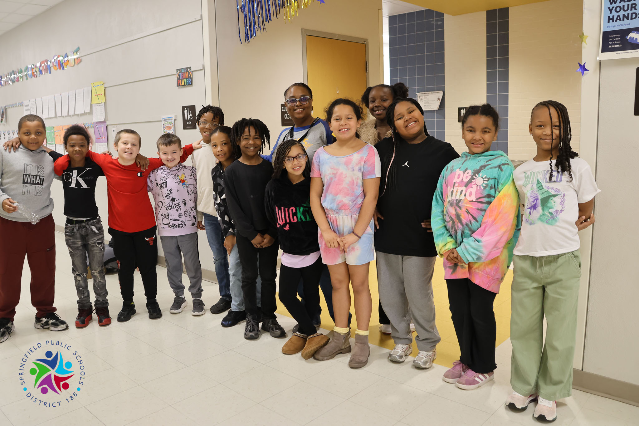 Students, in various colored tops and bottoms, stand in a hallway, smiling for a photograph.
