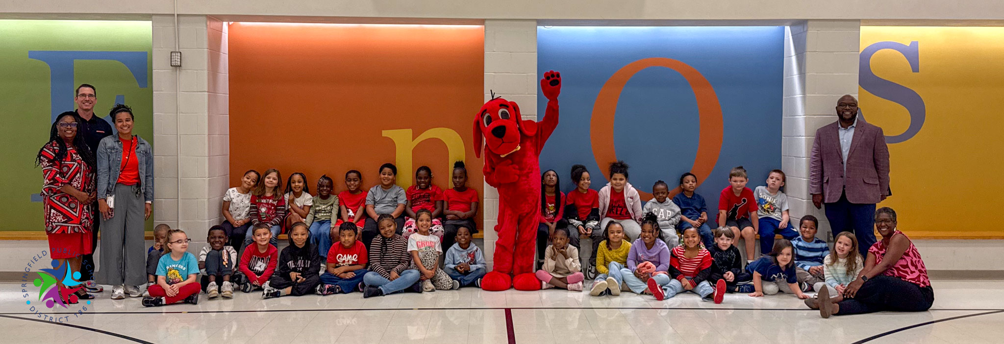 A group of children and adults pose for a photo with a red mascot in front of colorful letters.