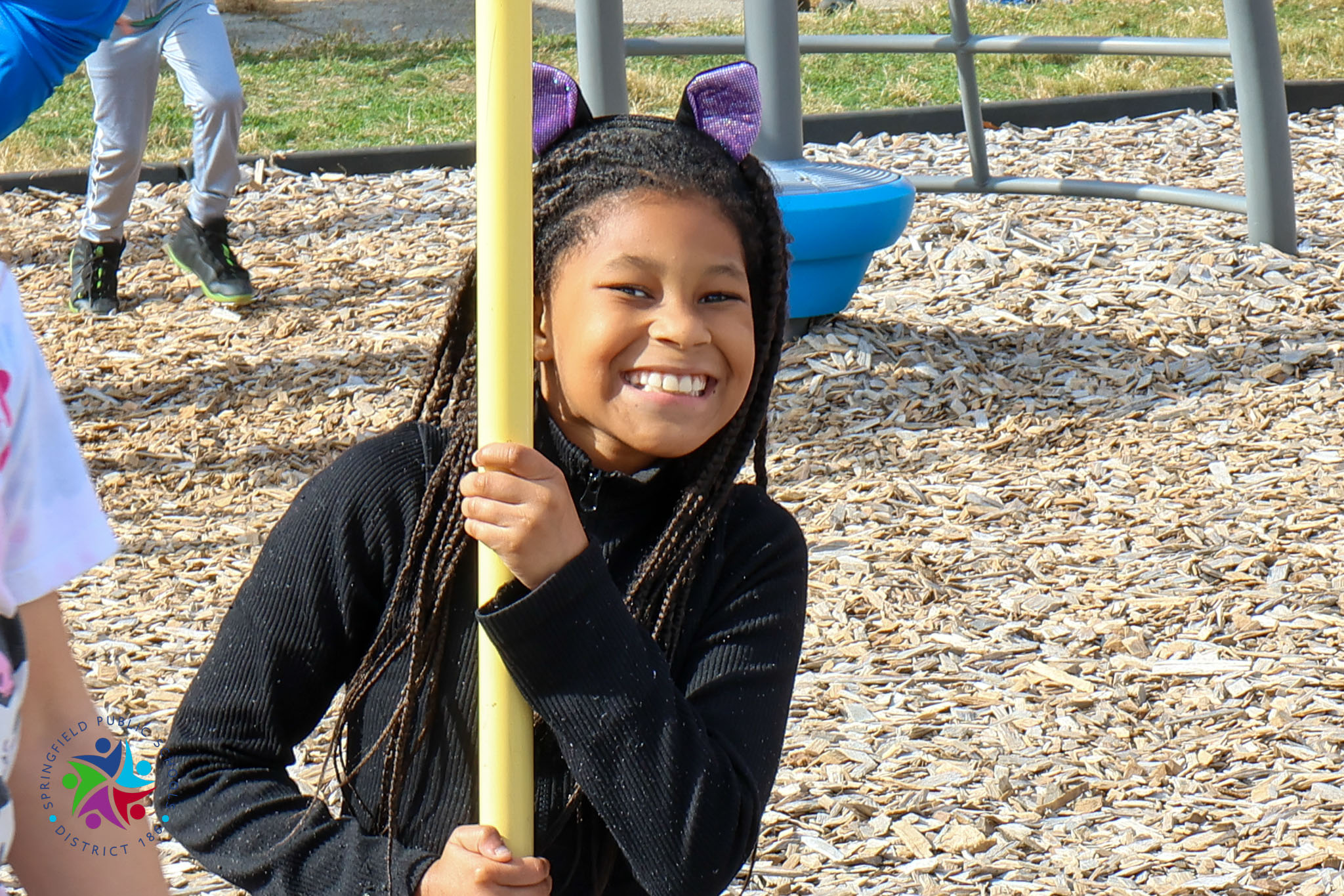 Child holds yellow pole with cat ears at playground. Child has long braids and wears a black top.