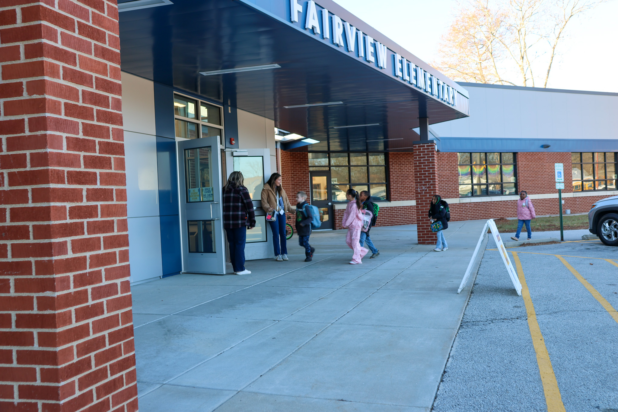 People are walking near the entrance of Fairview Elementary School. A brick wall is on the left.