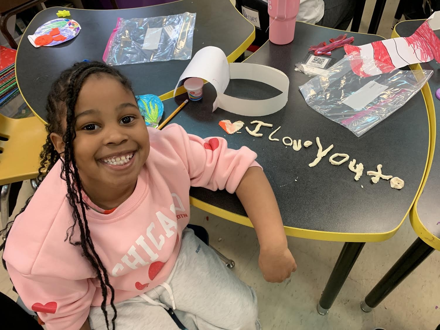 A smiling child sits at a table with craft materials, a pink shirt, and "I Love You" written on the table.