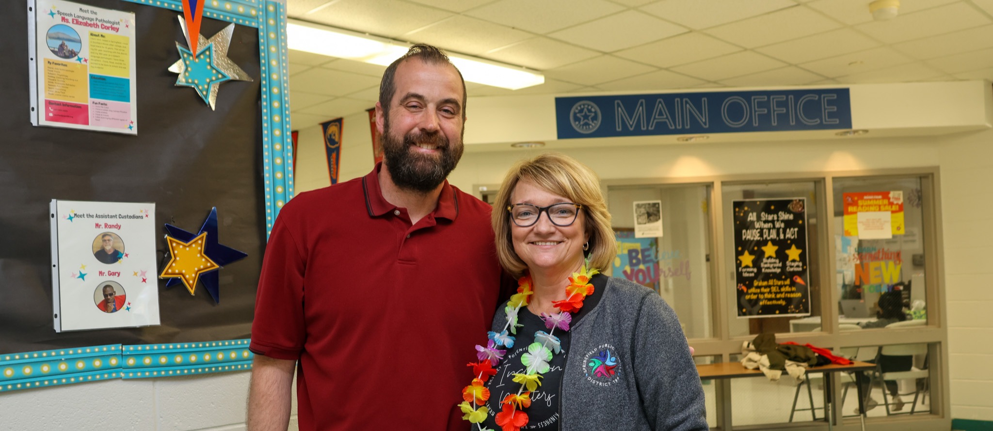 Man and woman standing in a hallway with a bulletin board behind them and a sign that reads "Main Office".
