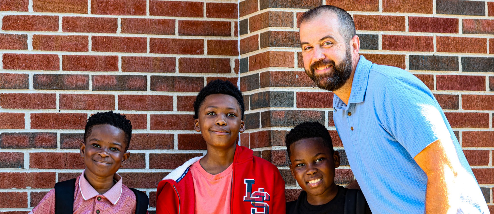 Four children stand near a brick wall. A man in a light blue shirt is smiling and leaning against the wall.