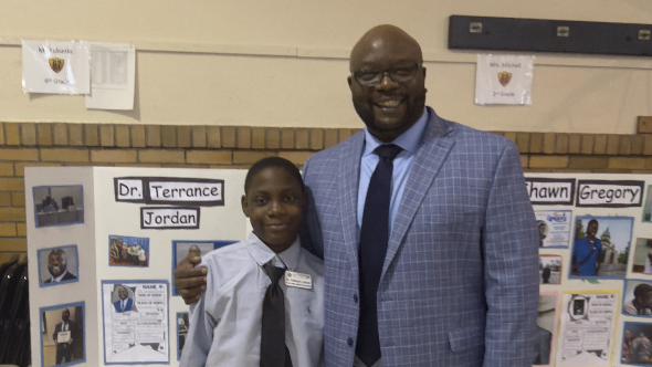 Man and boy in suits posing together in front of a bulletin board with photos.