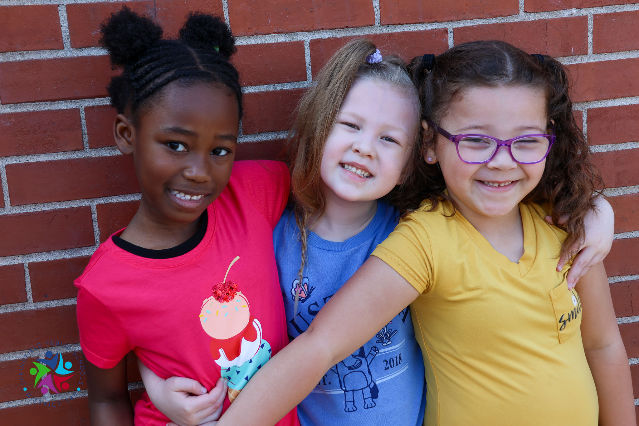 Three girls with different hair styles and glasses stand close, smiling for the camera. They wear colorful shirts.