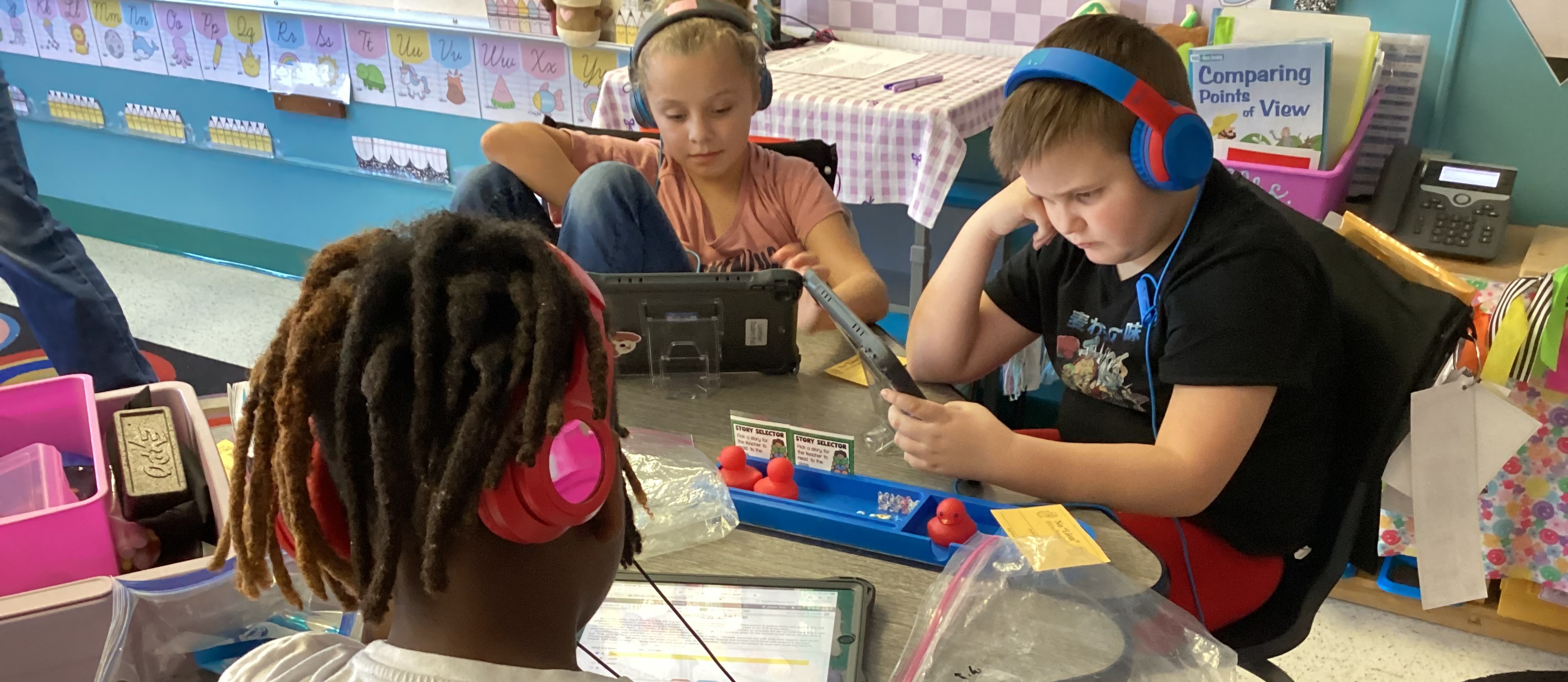 Two students with headphones use a tablet in a classroom with chairs and a table.