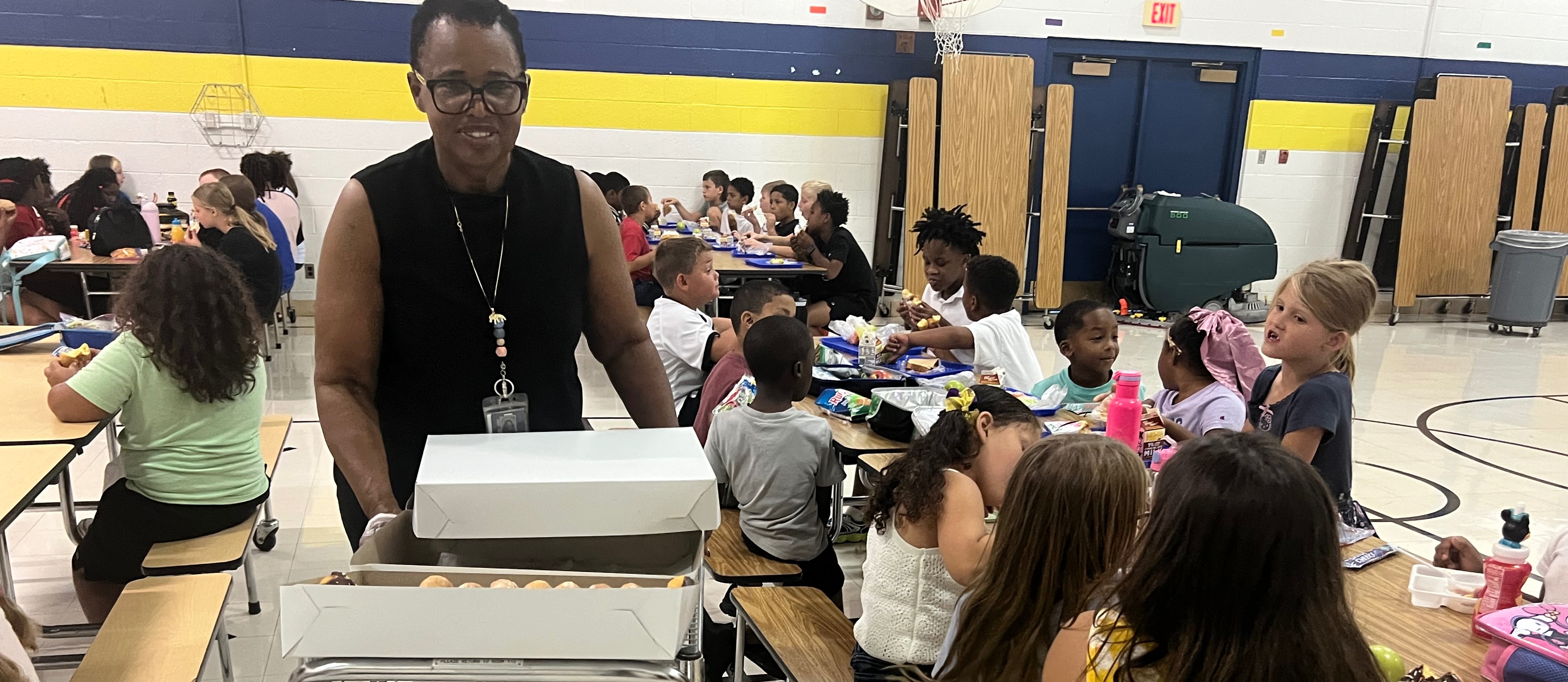 An adult stands in a cafeteria, overseeing children sitting at tables, engaged in activities. The room has yellow and blue walls.