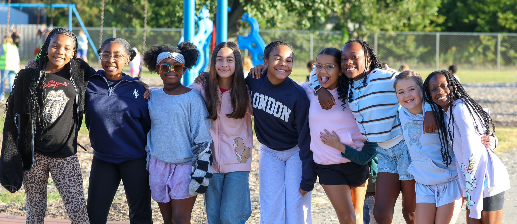 Nine girls and one boy stand and sit in a group outside, with trees and a playground behind them.