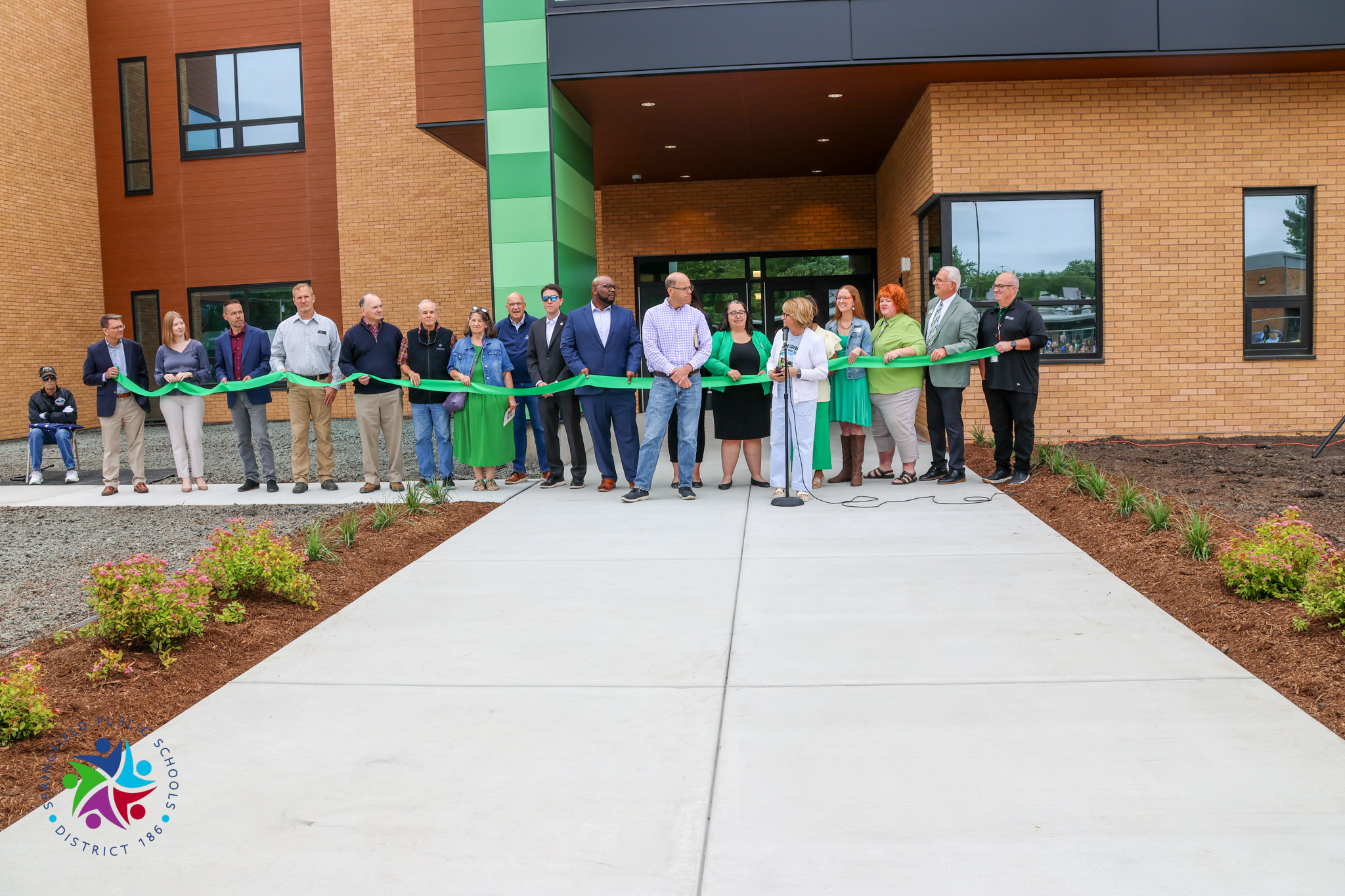 A group of people stands in front of a building holding a green ribbon for a ribbon cutting.