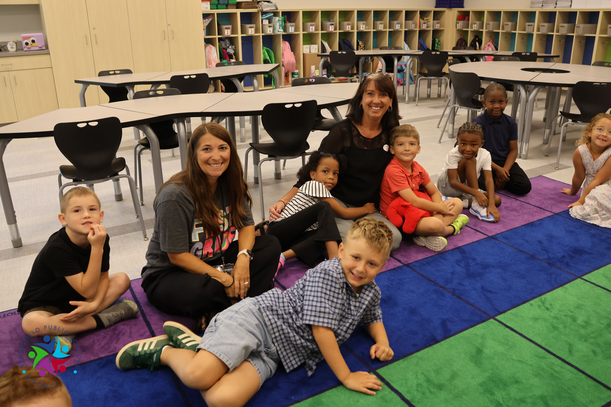A group of children and two women sit on a colorful carpet in a classroom with tables.