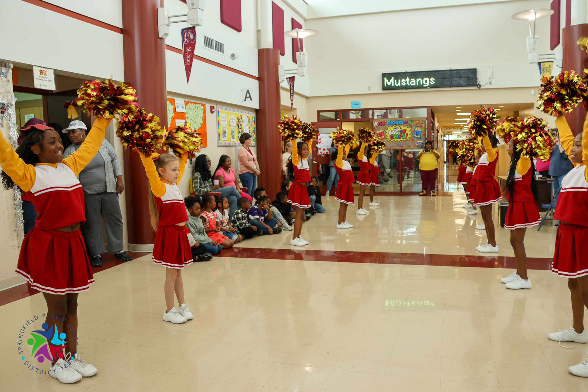 Cheerleaders in red and white uniforms perform in a school hallway, with spectators seated on the floor.