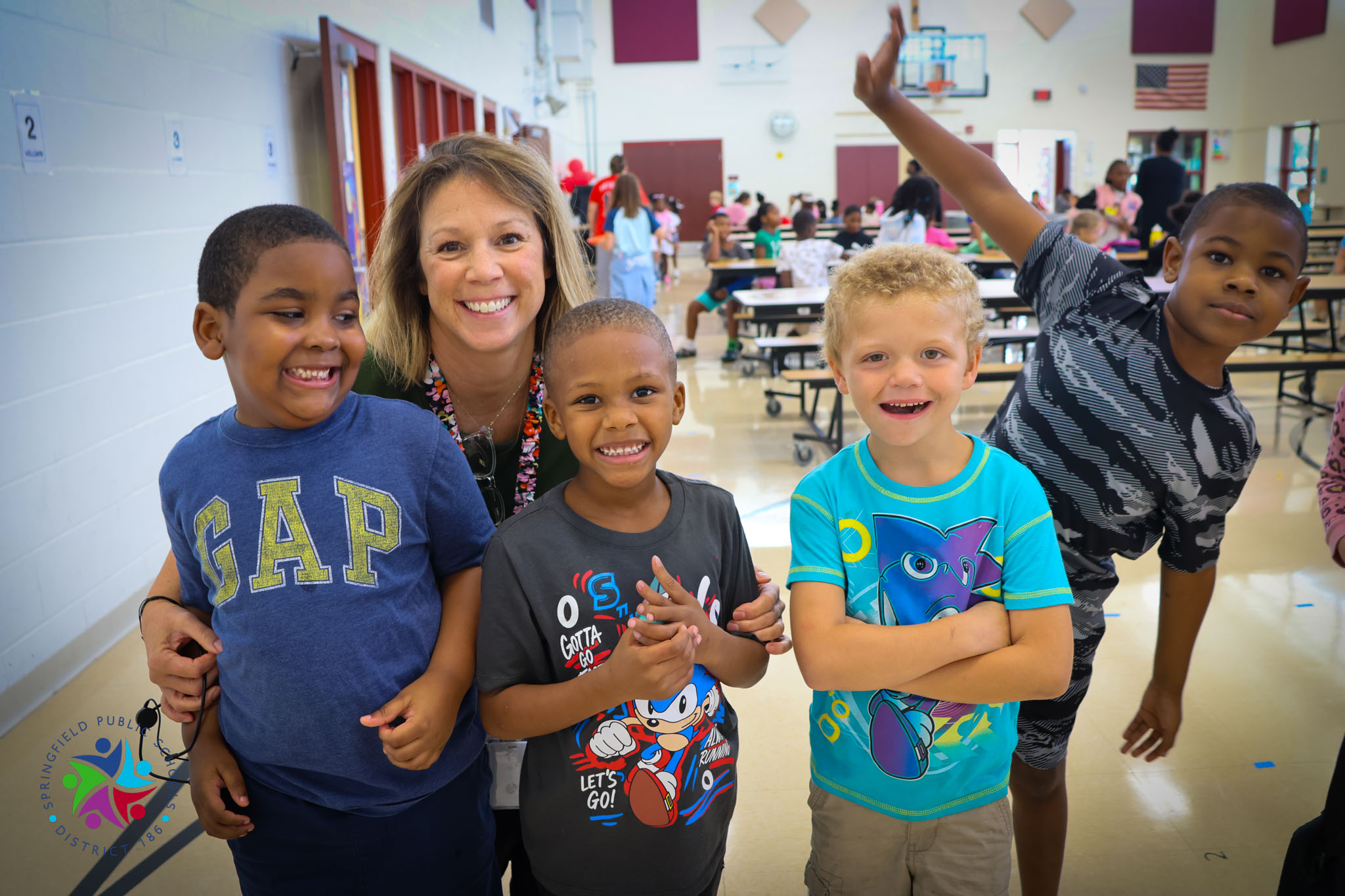 Woman stands with four smiling children in a cafeteria with tables and chairs in the background.