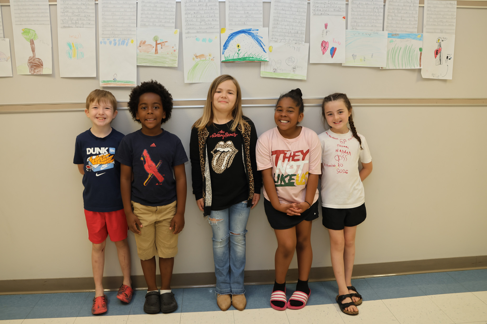 Five children stand together in front of a wall with drawings attached. They wear casual clothes.