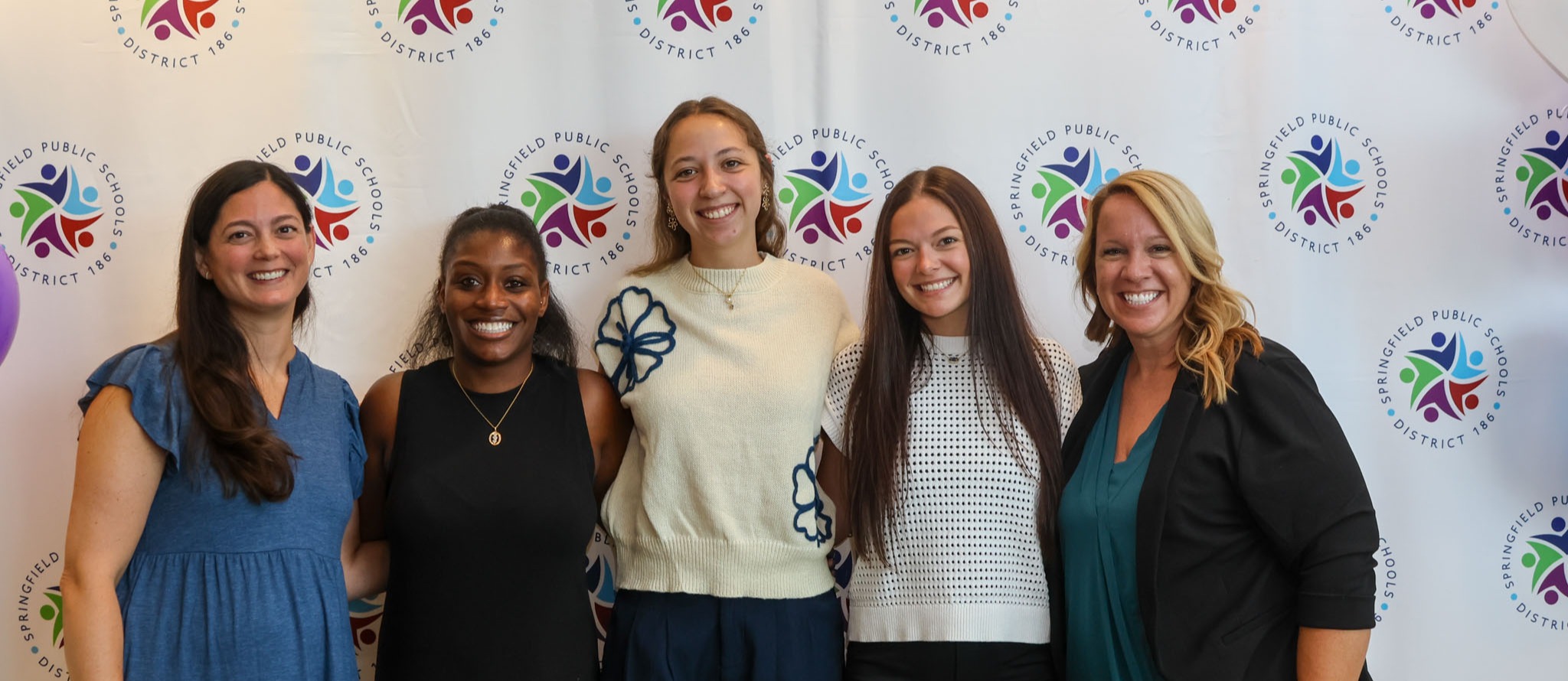 Five women stand next to each other in front of a wall with colorful logos and balloons.