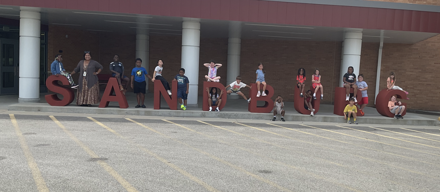 A group of individuals, some standing on letters, pose for a photo in front of a building.