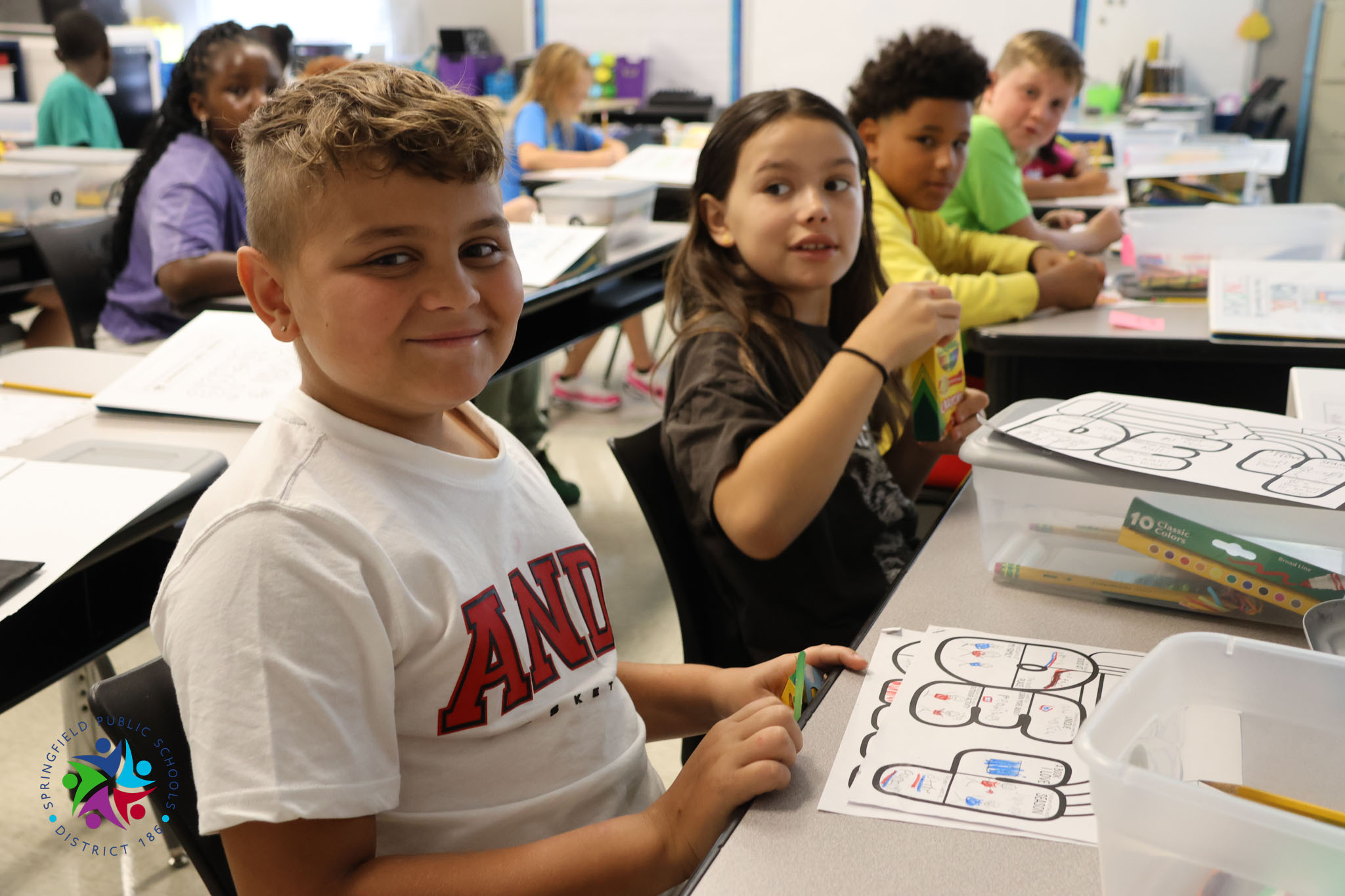 Children in a classroom. One boy smiles, another girl holds a book. Desks with papers, books, and a plastic container.