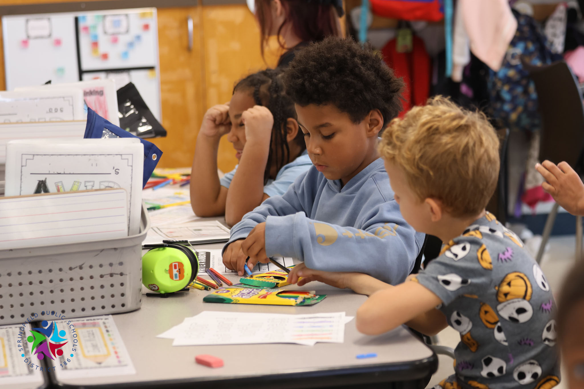 Three children sitting at a table, using scissors and markers to cut and color papers.