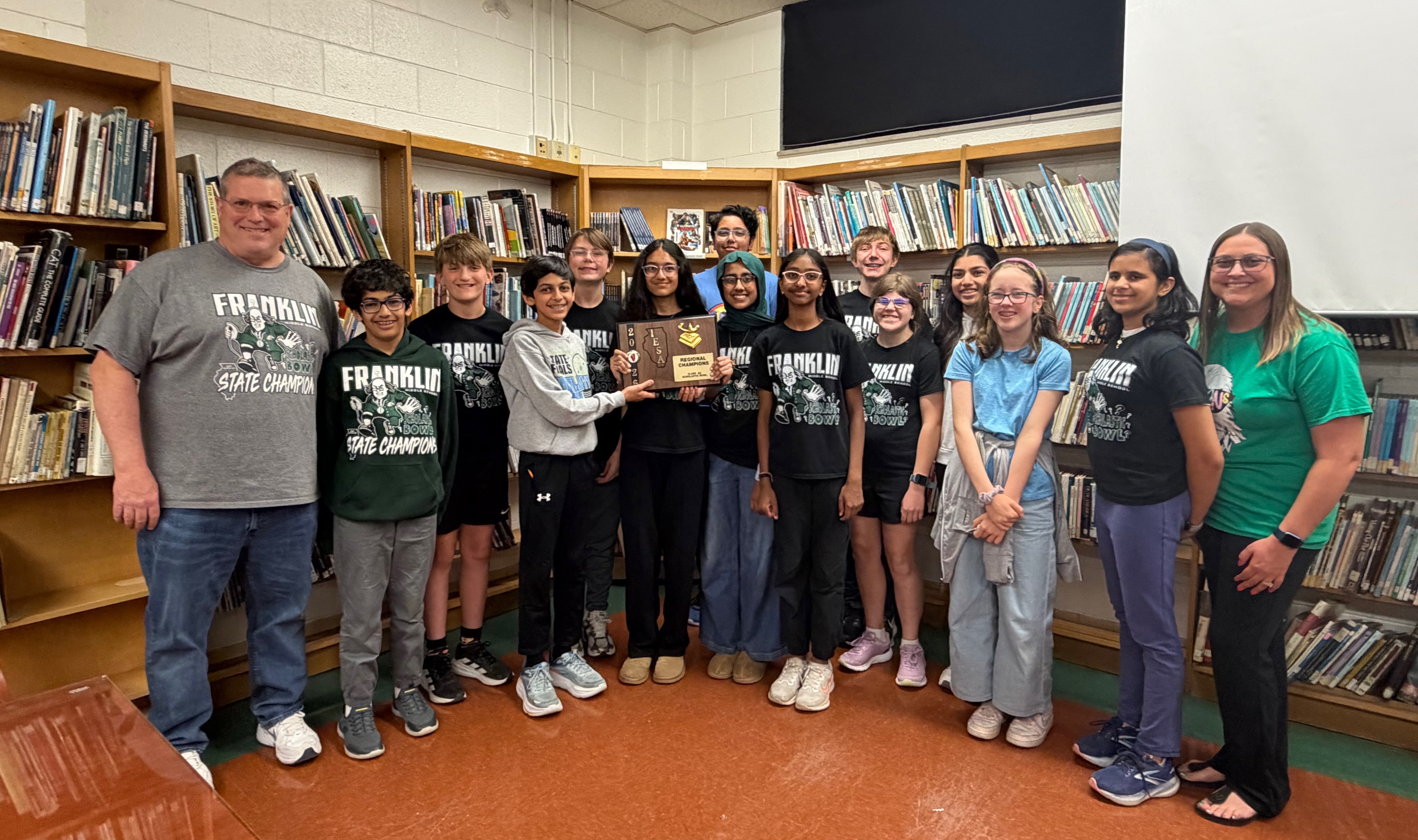 A group of students and adults pose with a trophy in a library filled with bookshelves.