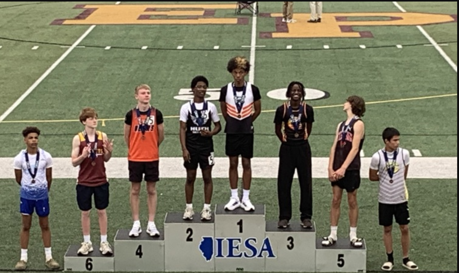 Five young athletes stand on a podium, medals around their necks, on a football field.