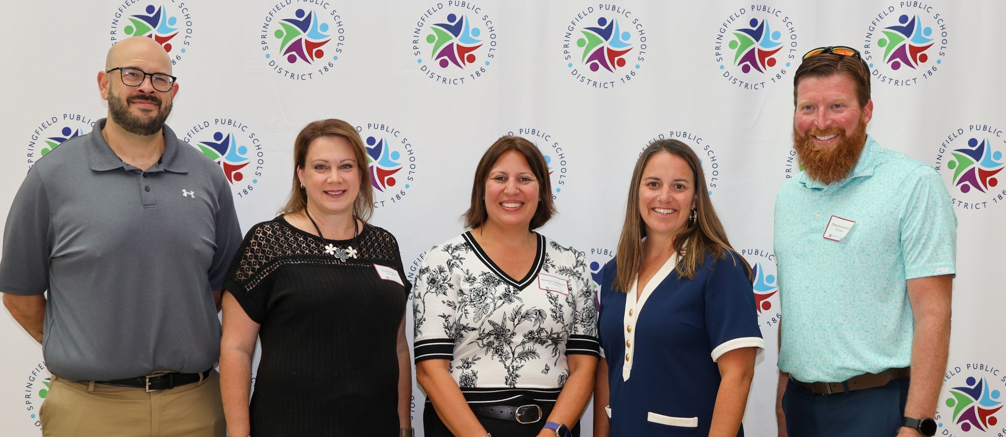 Five people stand in front of a backdrop with logos and colors. They are all smiling.