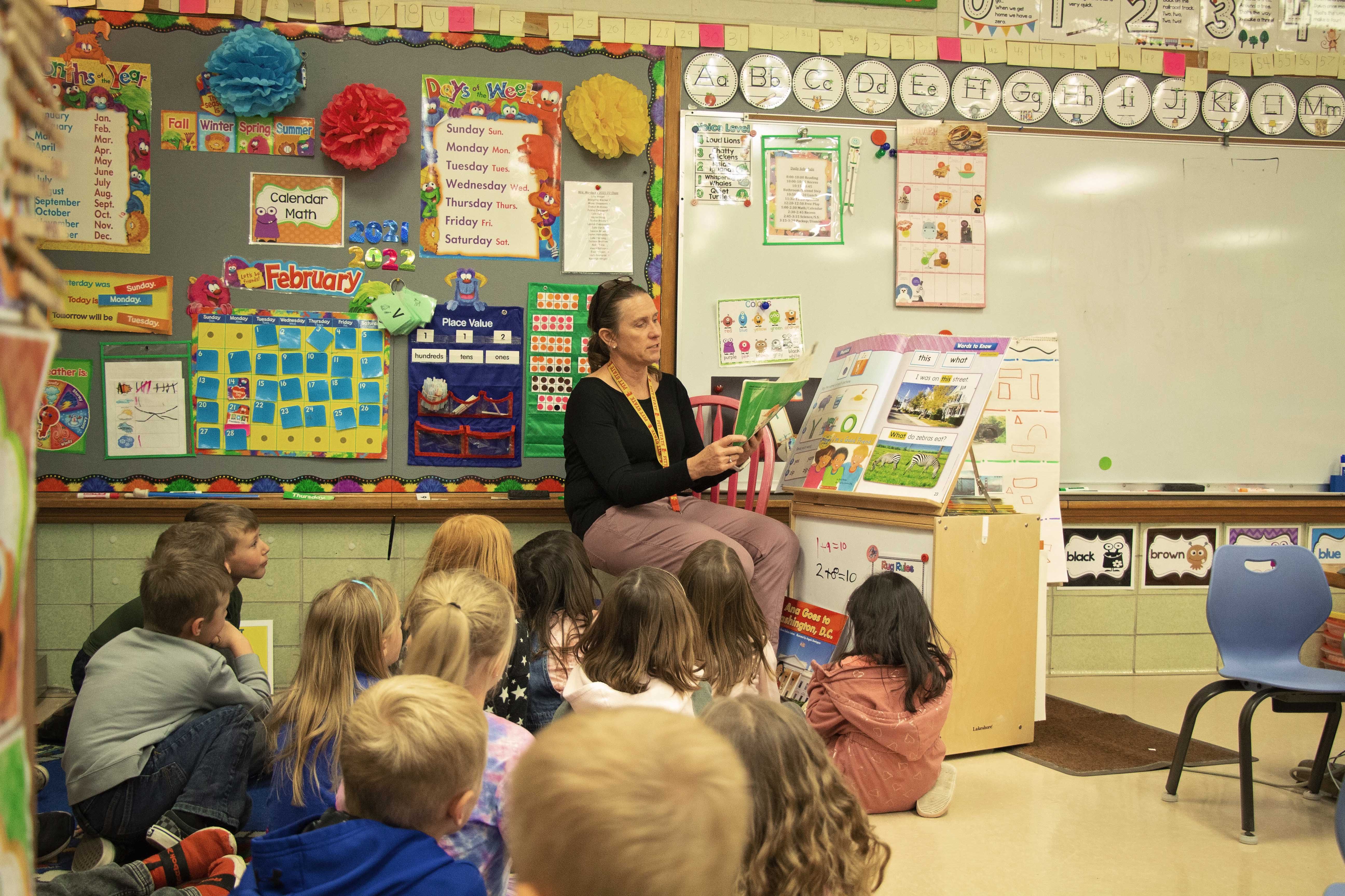 Kindergarten teacher reading book in front of students in a classroom.