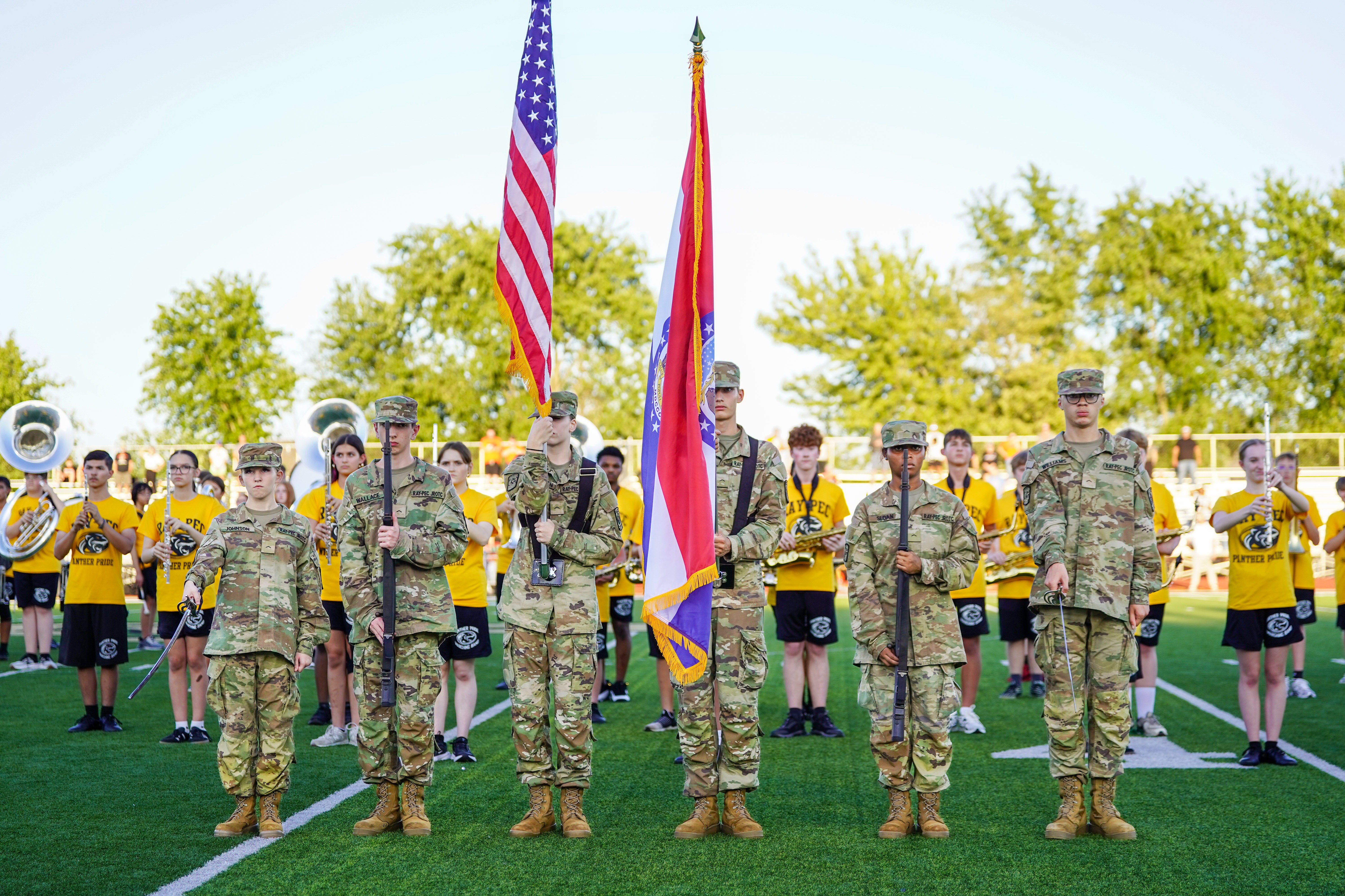 Students in army uniforms stand on a football field with flags with a marching band in the background.