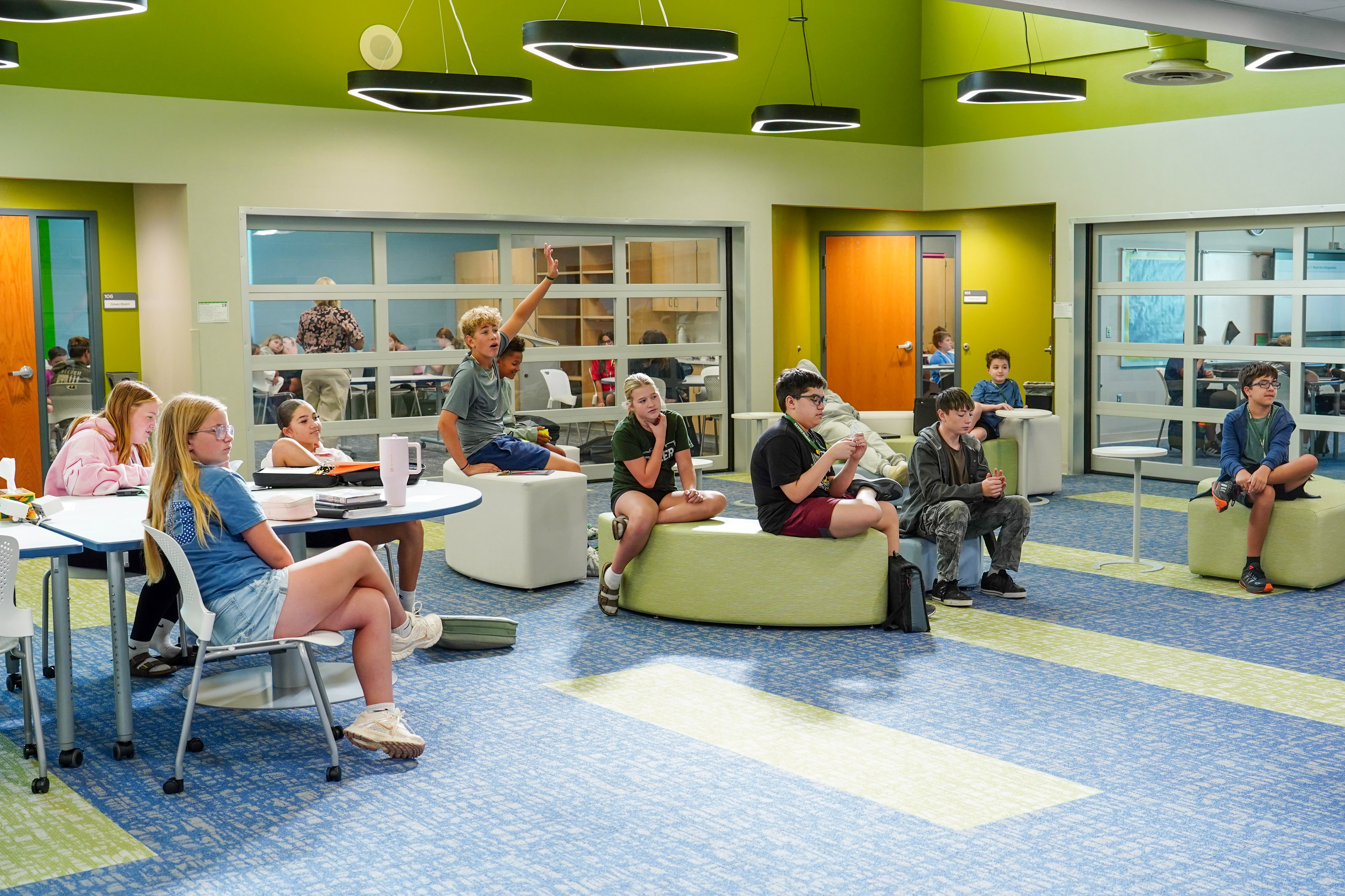 A group of students sitting around an open classroom listening to the front of the room.
