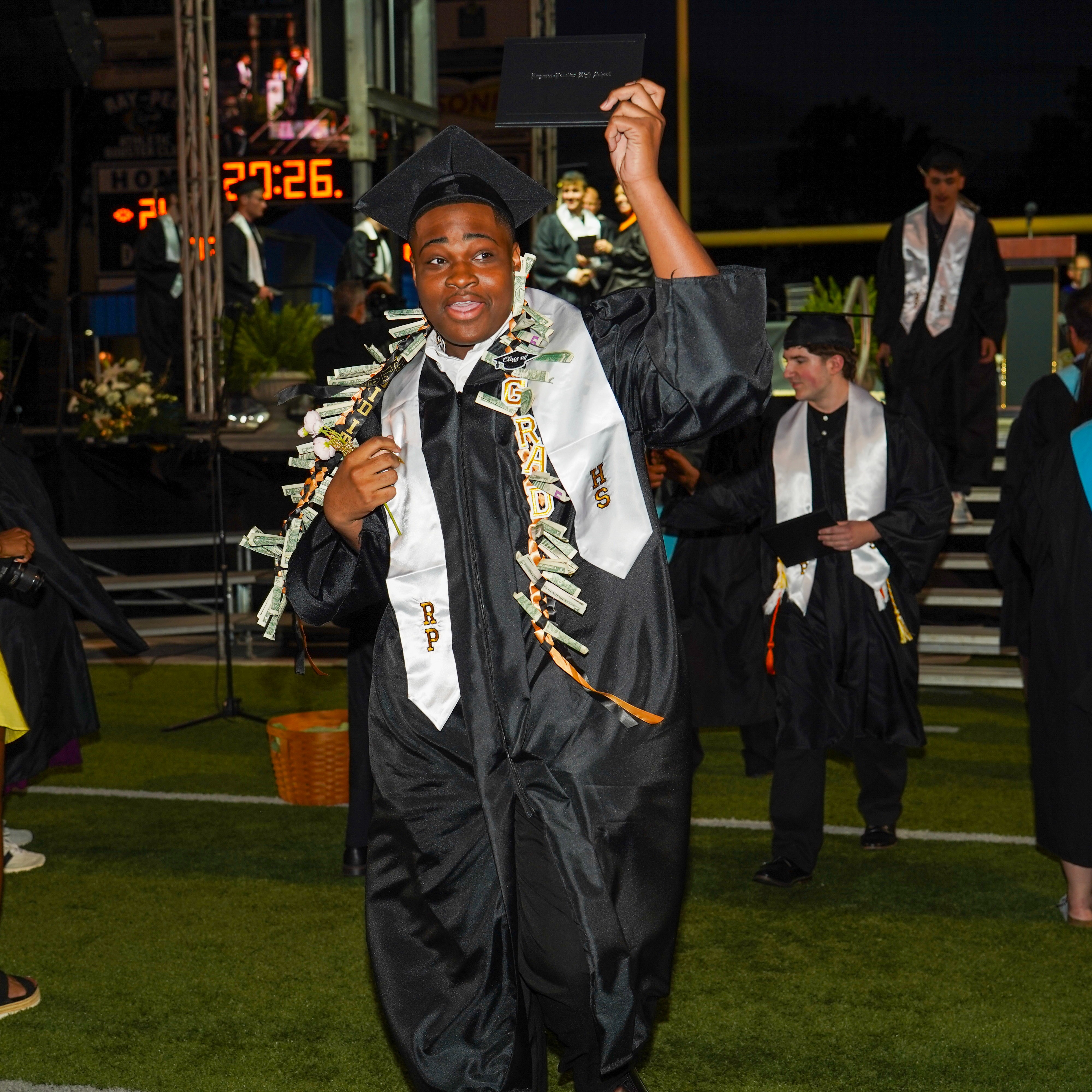 A graduate in a black robe and cap holds his diploma up in celebration.