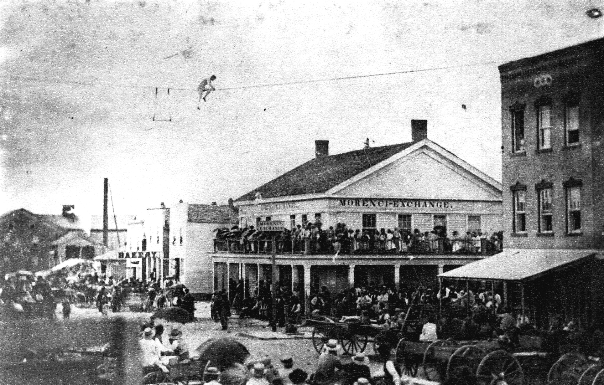 Centennial Celebration July 4, 1876 at corner of West Main and North Streets.  Abraham Babcock walking tight-wire from Rorick’s Hardware (Bank Bldg. site) to former Mace Bldg. N.E. corner.  Morenci Exchange sign and village pump in street.  Bakery and the grist mill in distance.