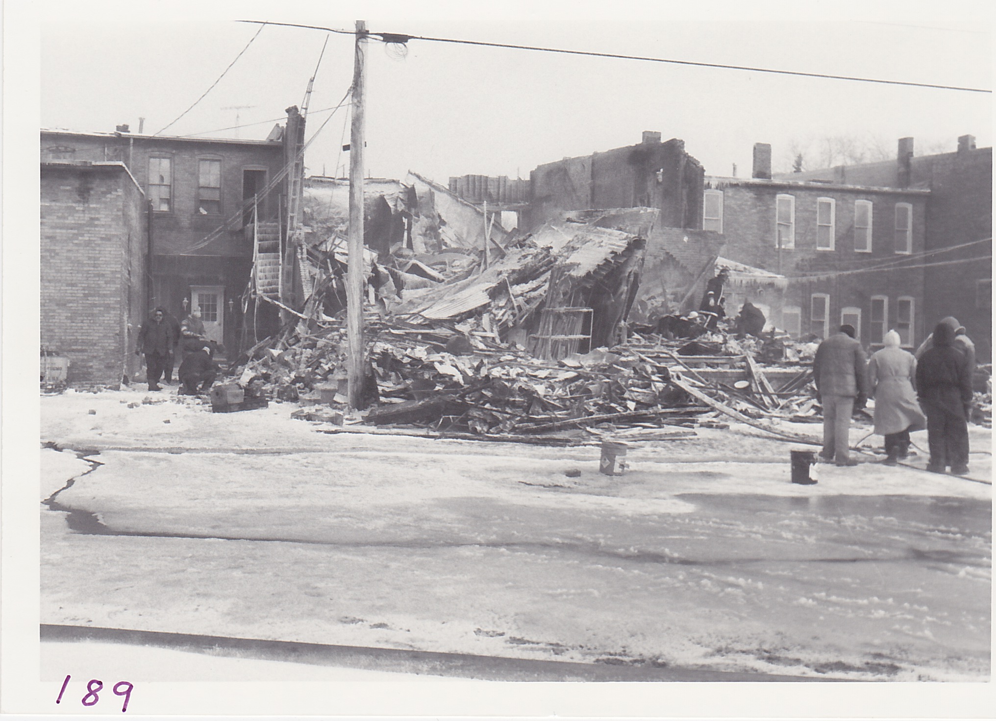 A rear view of damaged Gamble Store Building.  Firemen can be seen looking for store records among debris.