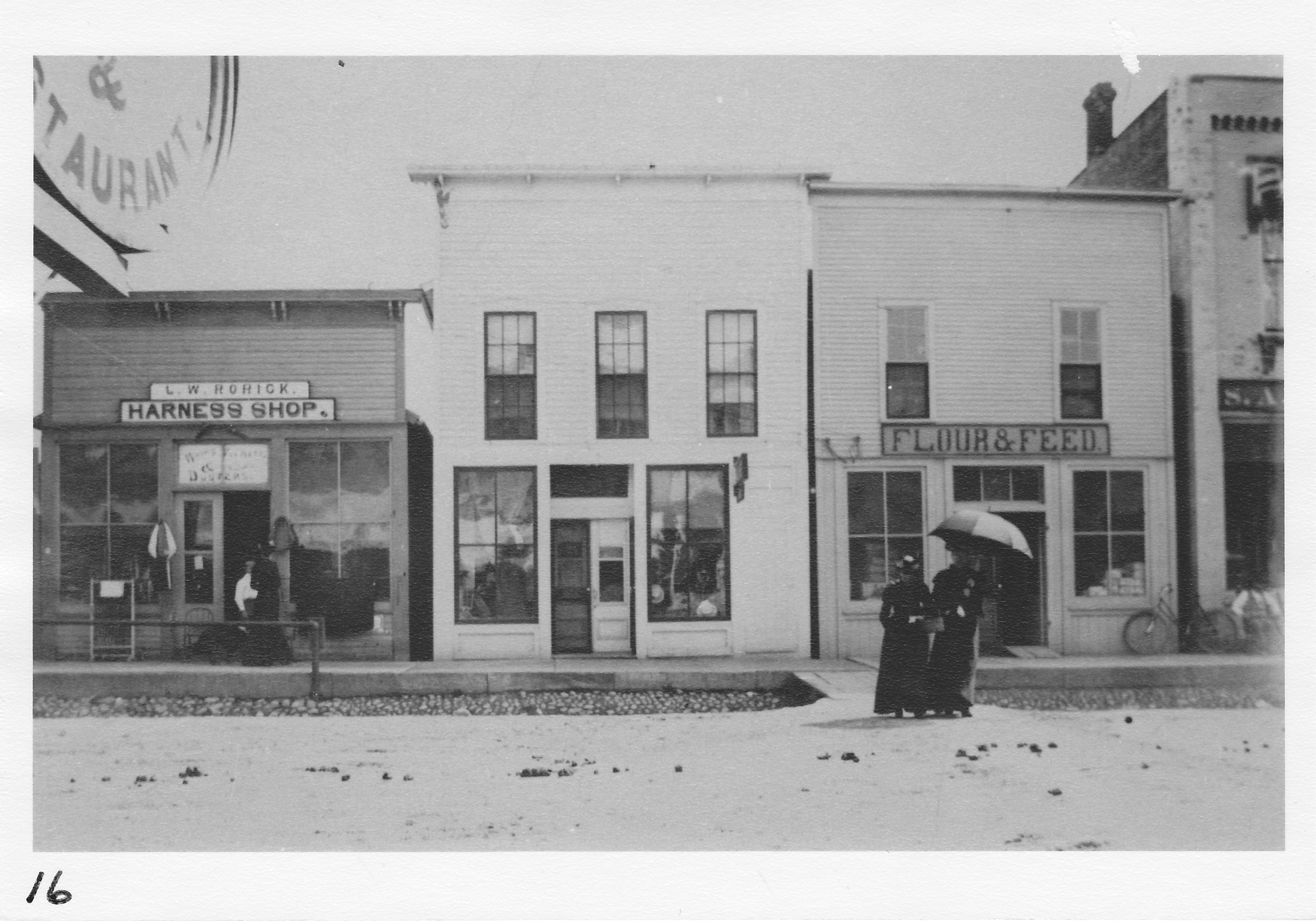 West Main Street, south side.  Mrs. Ellen Oldfield and her sister crossing the street.  Other buildings:  Lewis R. Rorick Harness Shop; Killin Hat Shop; Flour and Feed Shop (later Brasher dentist office & Rex Theater ), Scofield’s Store, extreme right.