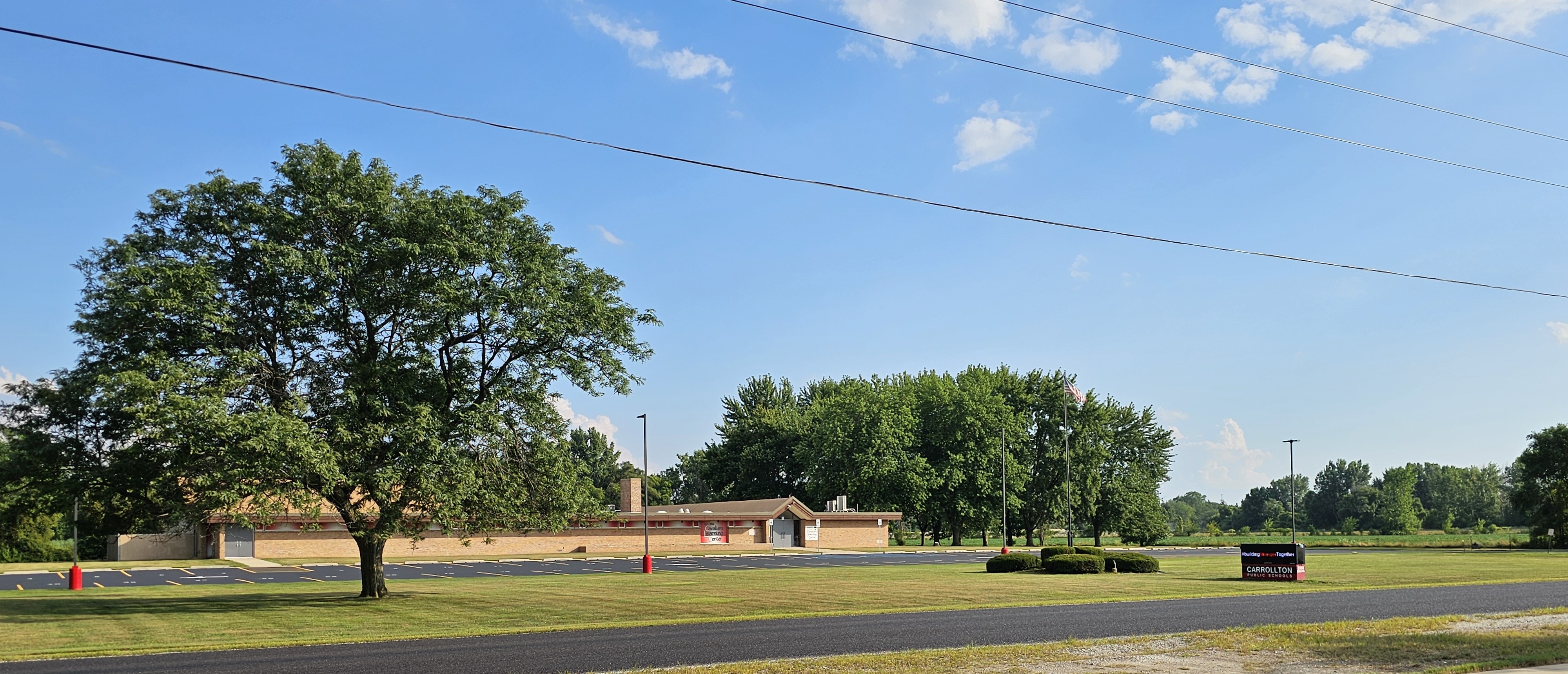 Wide view of a Carrollton Public Schools campus on a sunny day, showing a single-story brick building behind a large lawn with mature trees. An American flag flies near trimmed bushes in the center of the lawn. The school’s digital sign, reading “Carrollton Public Schools,” stands on the right side of the image. The sky is bright blue with scattered clouds, and power lines run across the top of the scene.