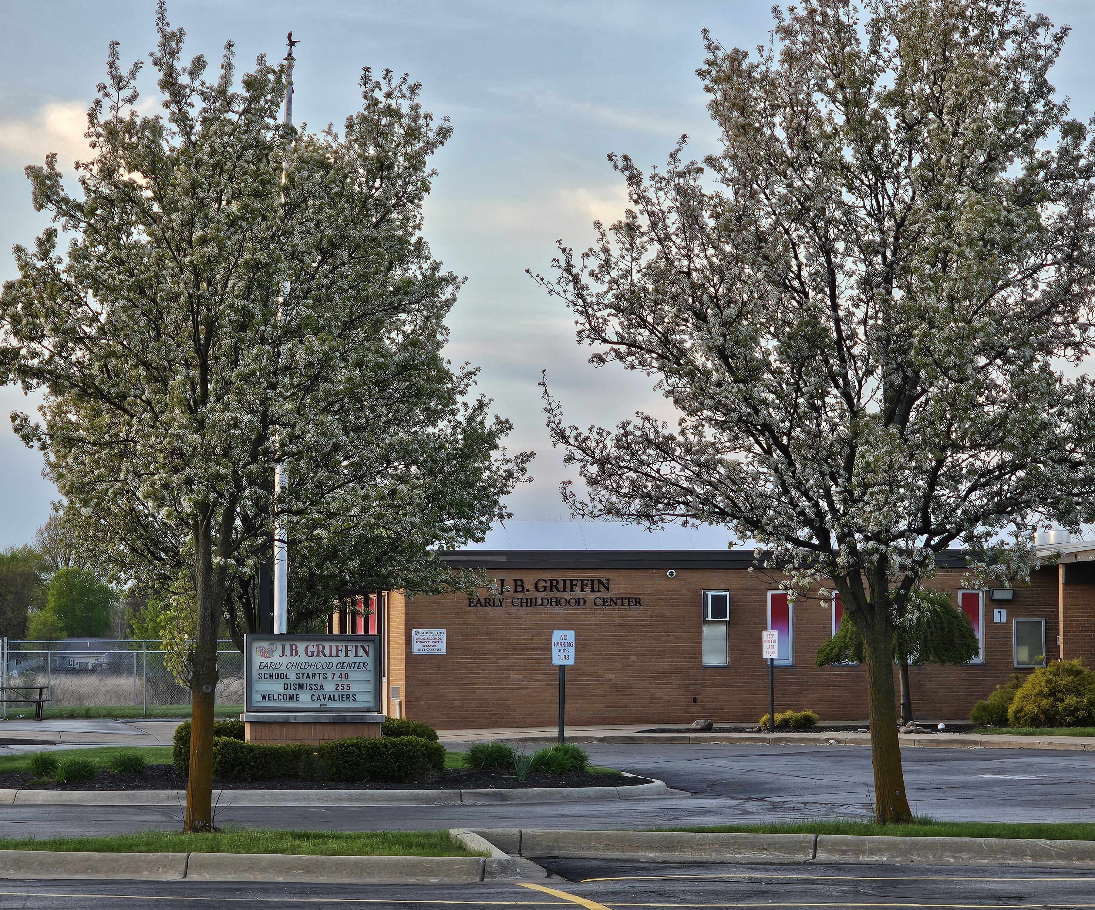 Exterior view of J.B. Griffin Early Childhood Center at Carrollton Public Schools during springtime. Two large flowering trees frame the front of the brick building, with the school sign visible near the entrance reading “J.B. Griffin Early Childhood Center.” The sky is cloudy and the parking lot and landscaping are visible in the foreground.