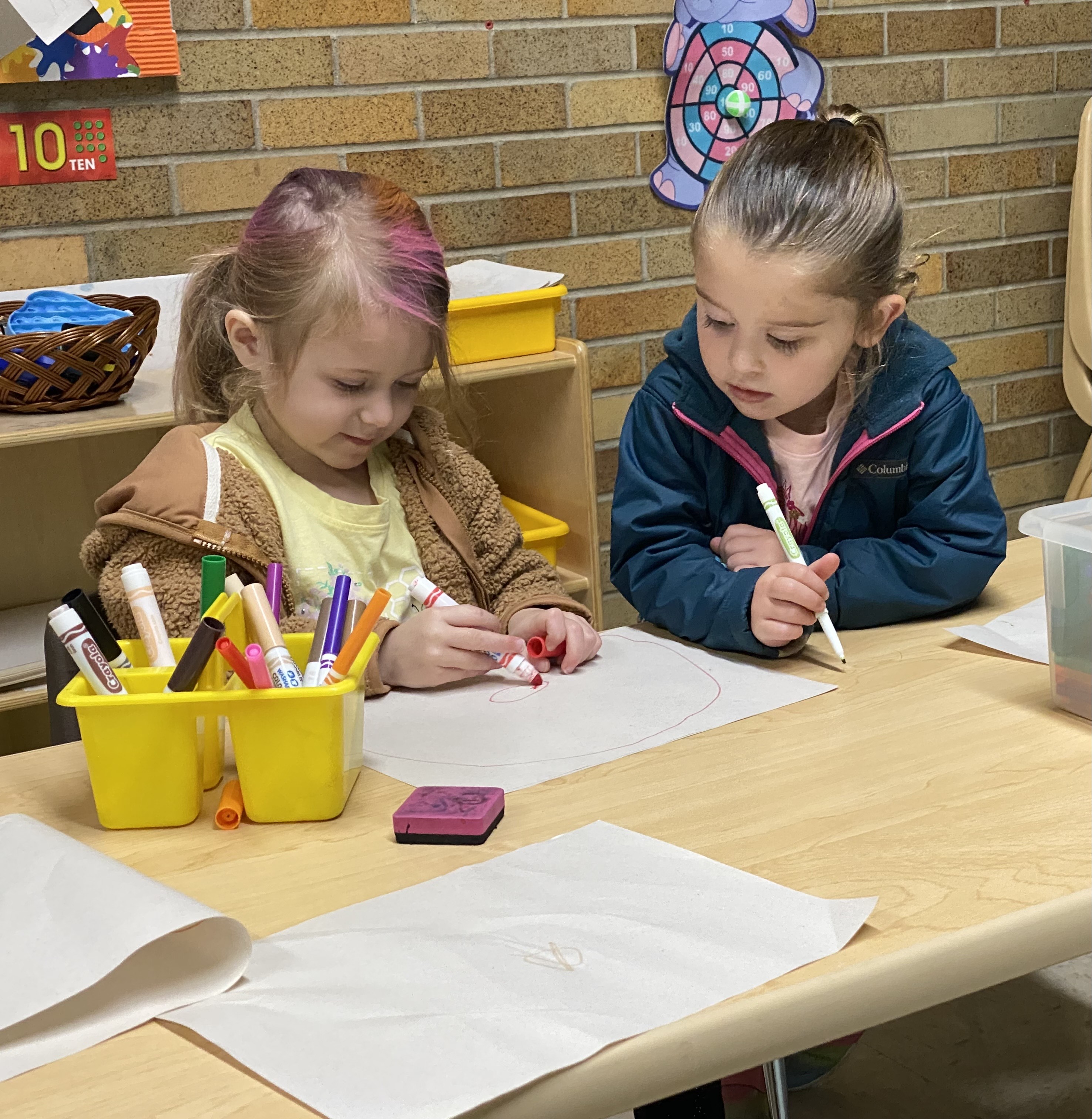 two girls drawing with crayons on white paper at table