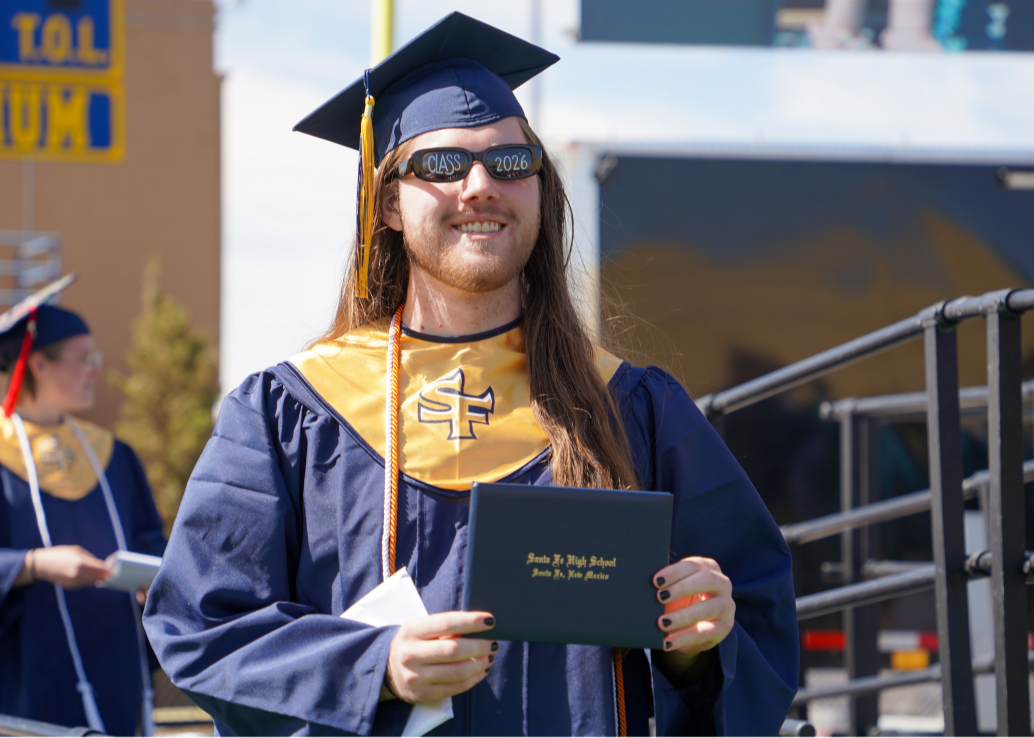 A person in a blue gown and hat smiles while holding a certificate at an outdoor event.