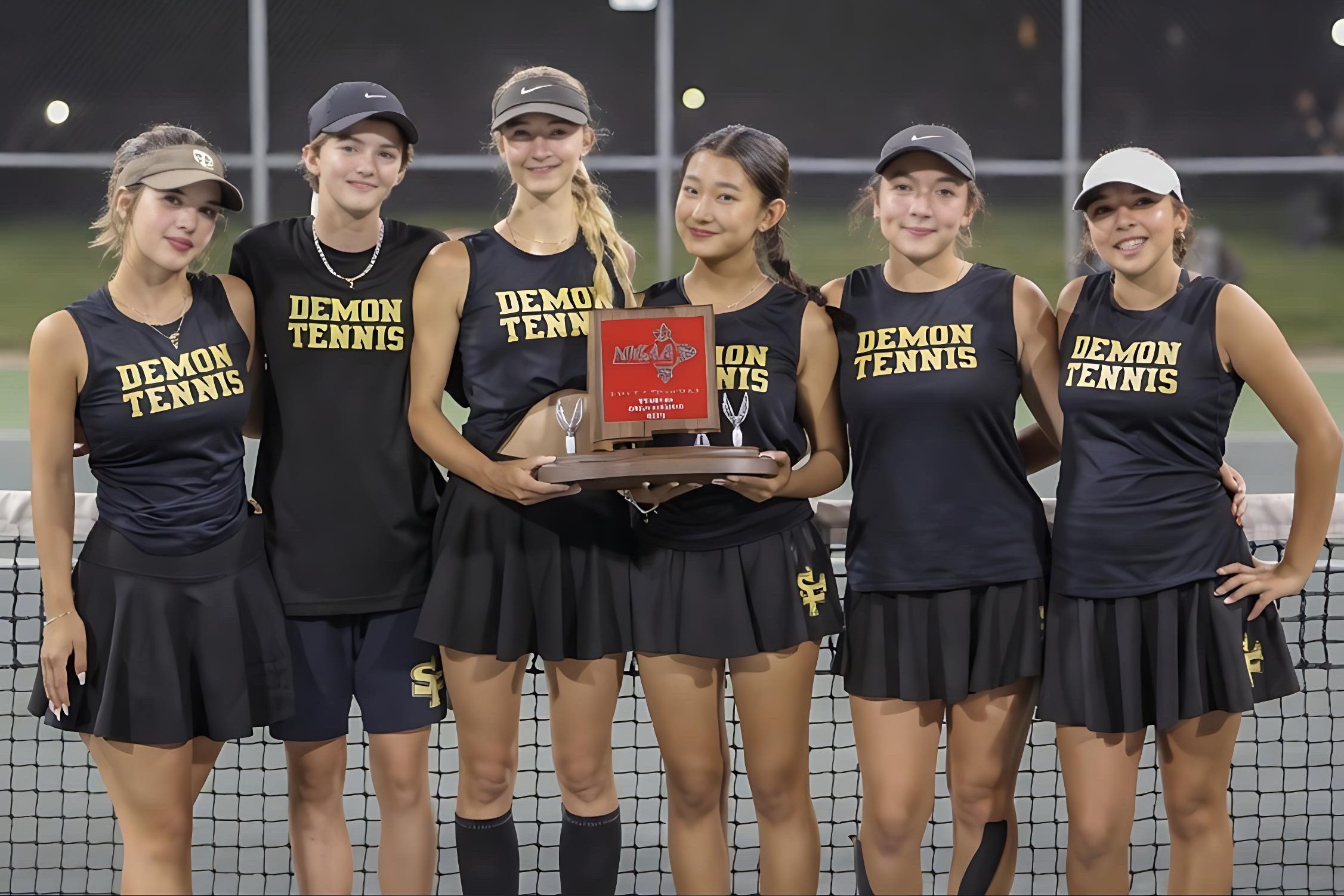 Six female tennis players pose for a photo, holding a trophy, wearing matching black uniforms. They stand on a tennis court.