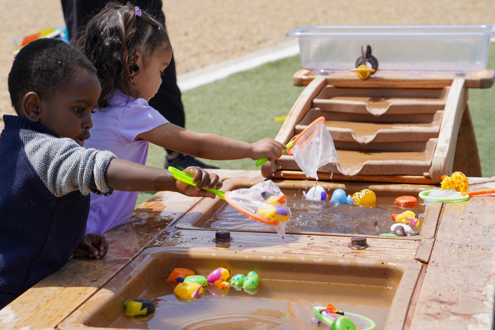 Two young children are playing with water and toys in a sandbox. One child holds a net.
