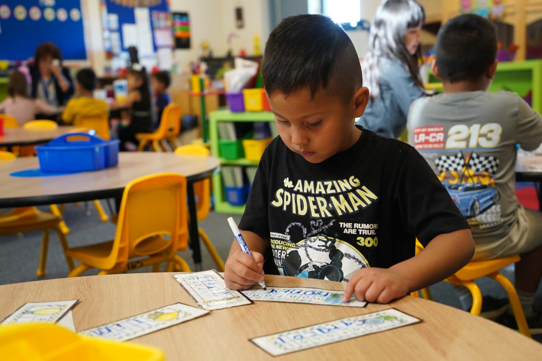 A child in a Spiderman t-shirt sits at a table, writing on cards. Other children are seated behind him.