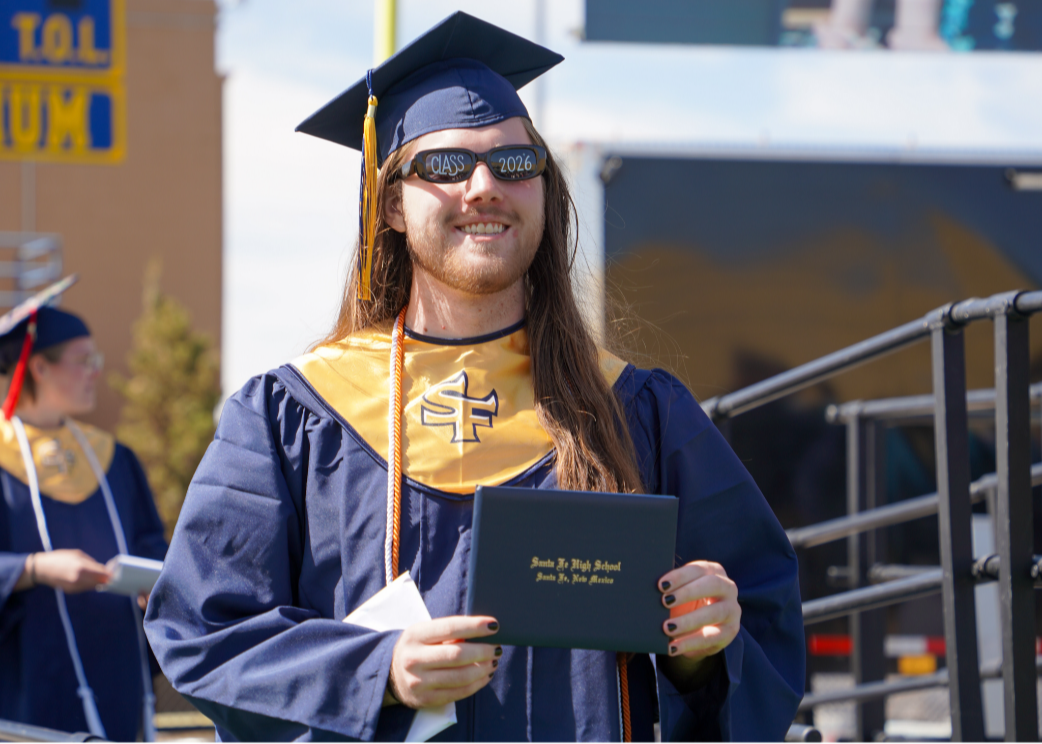 A person in a blue gown and hat smiles while holding a certificate at an outdoor event.