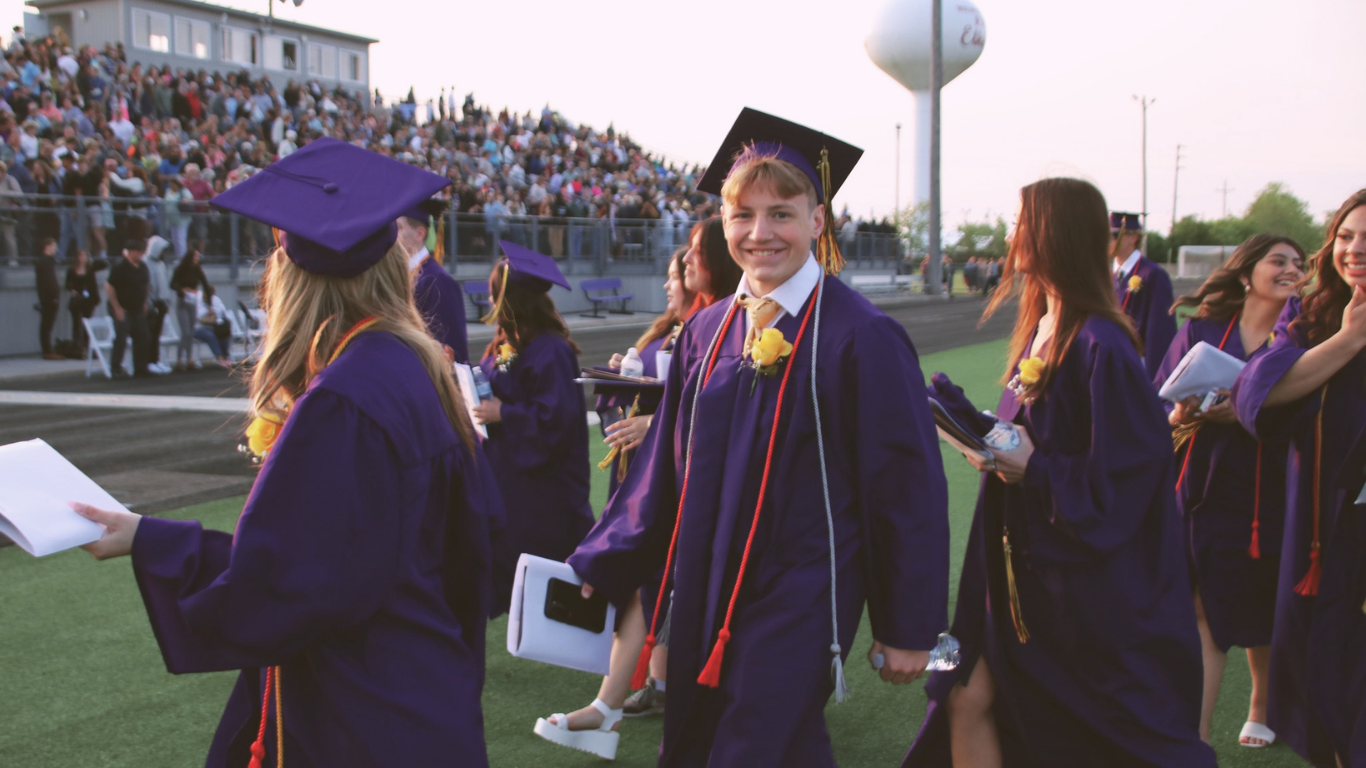 Graduates in caps and gowns walk on a field; one holds a book, another smiles. Behind them, an audience sits.
