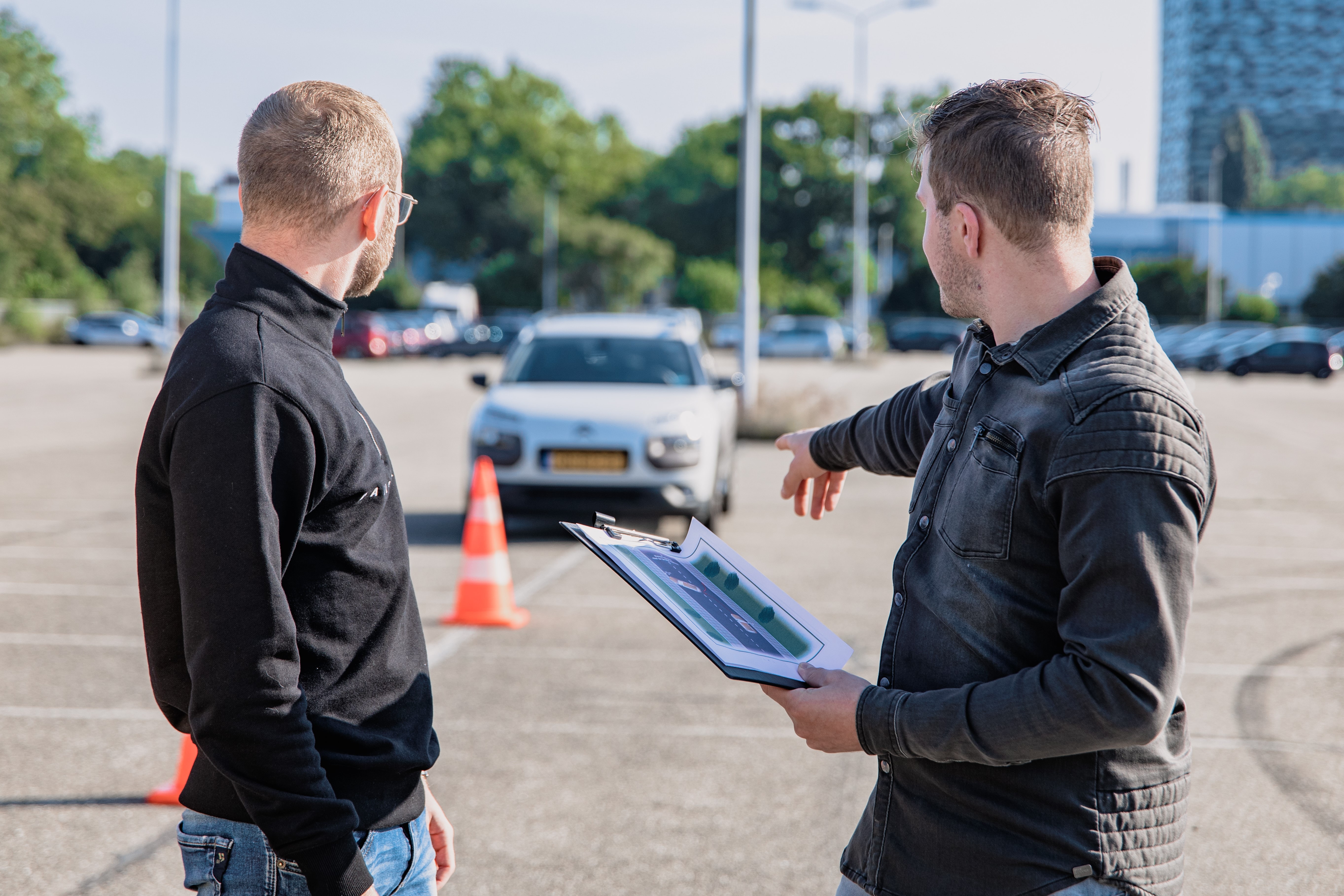 photo of two men pointing behind them, toward a car and an orange traffic cone.