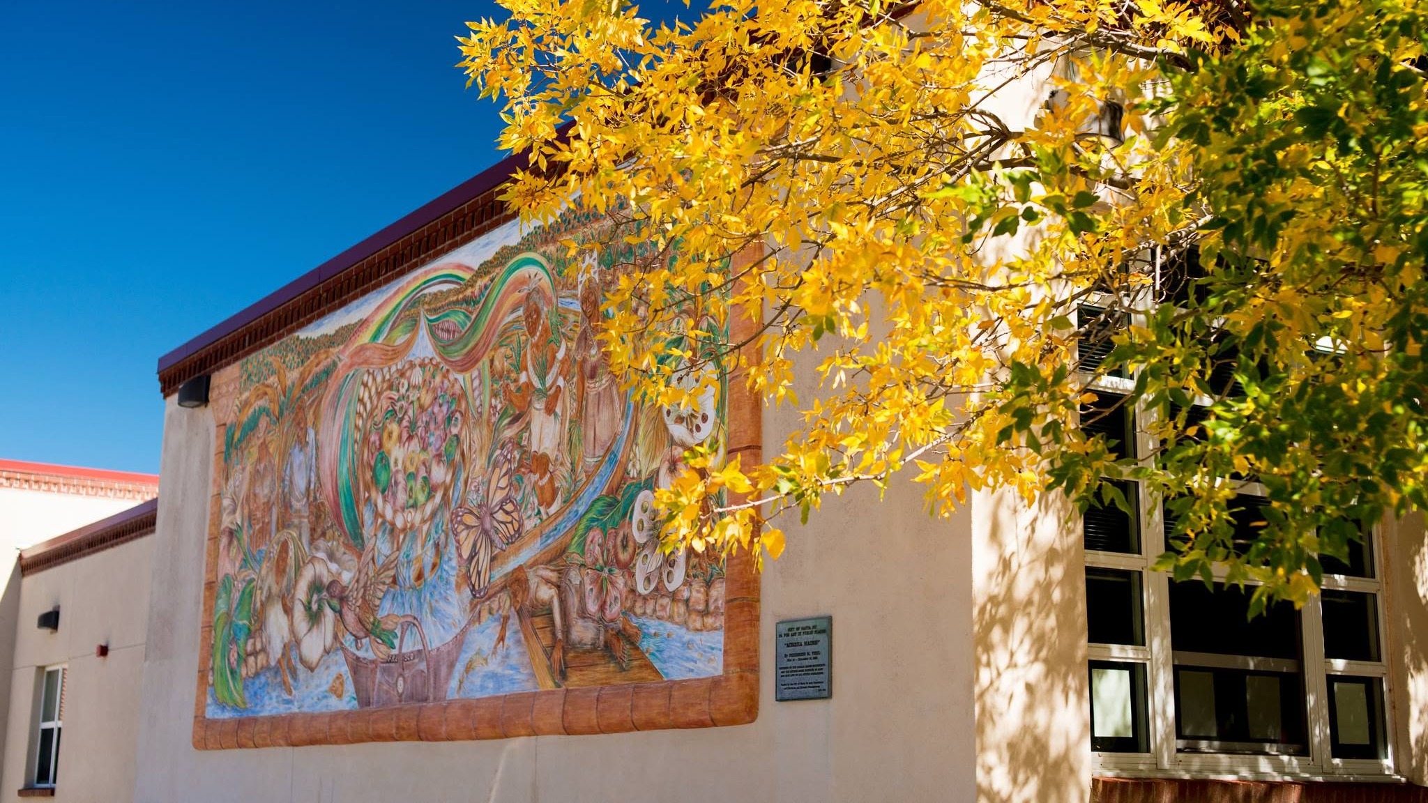 A mural adorns a white building wall, vibrant colors and designs. Yellow tree leaves surround the mural. Windows line the building.
