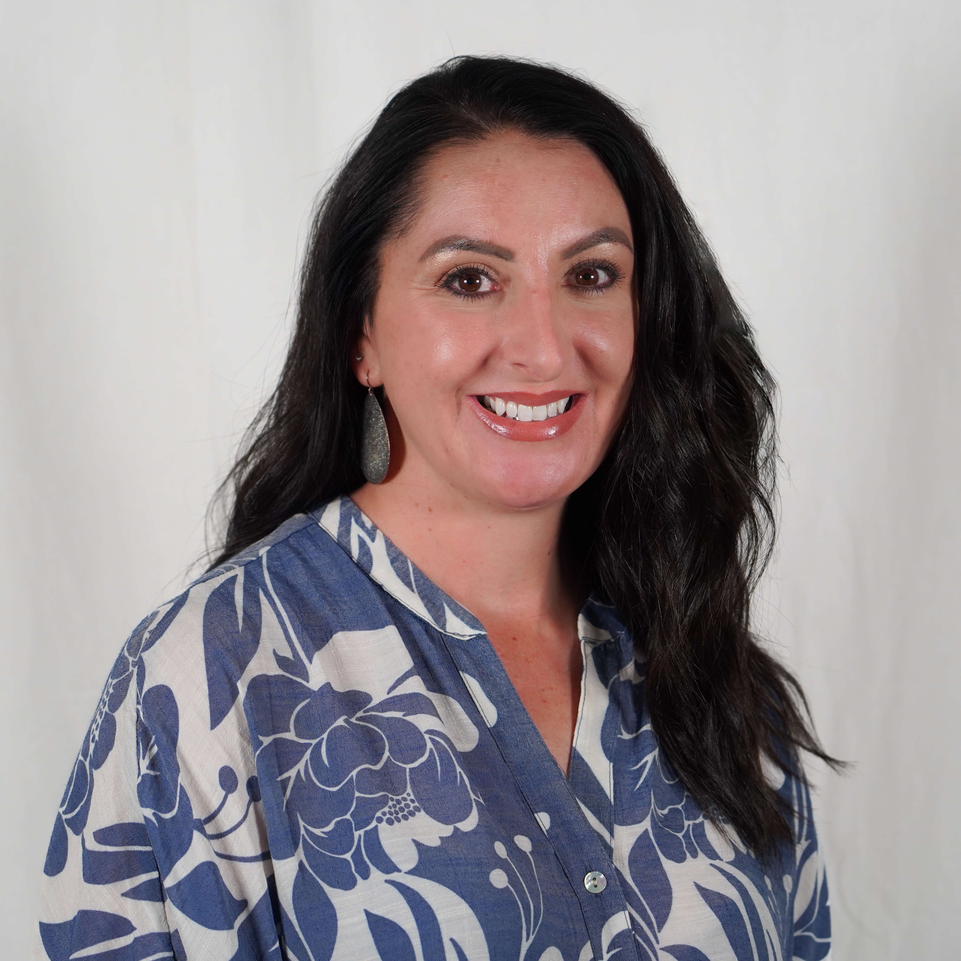 Woman with long black hair wearing a blue and white blouse with a flower pattern, smiling, standing against a white backdrop.