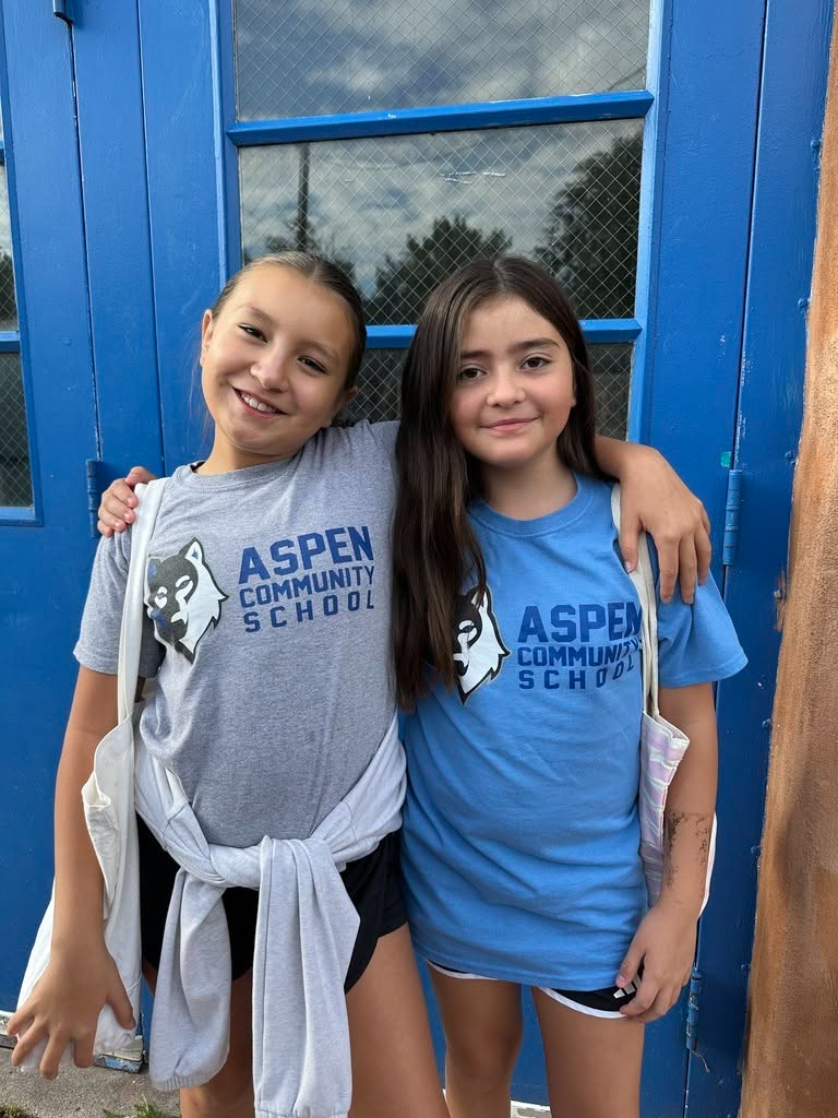 Two girls stand with arms around each other outside a blue door. They wear blue and gray T-shirts with "Aspen Community School" written on them.
