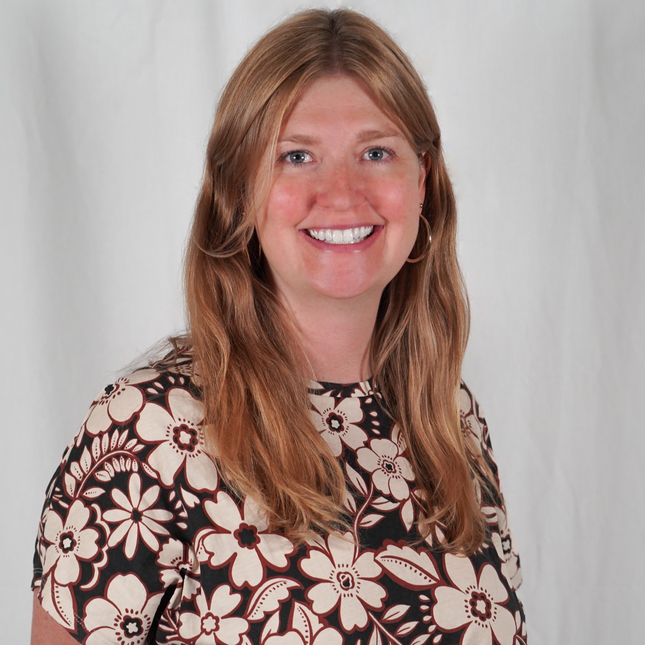 A woman with blonde hair wearing a floral blouse, standing in front of a white backdrop, smiling.